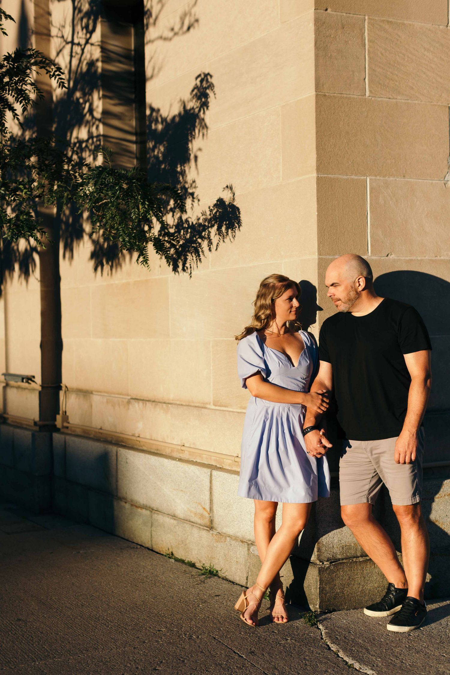Couple in casual attire poses playfully against beige wall during golden hour photoshoot.