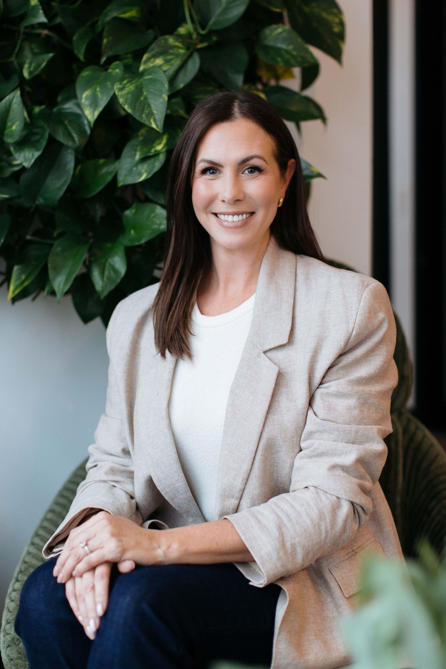 Professional headshot against leafy background wearing neutral blazer.