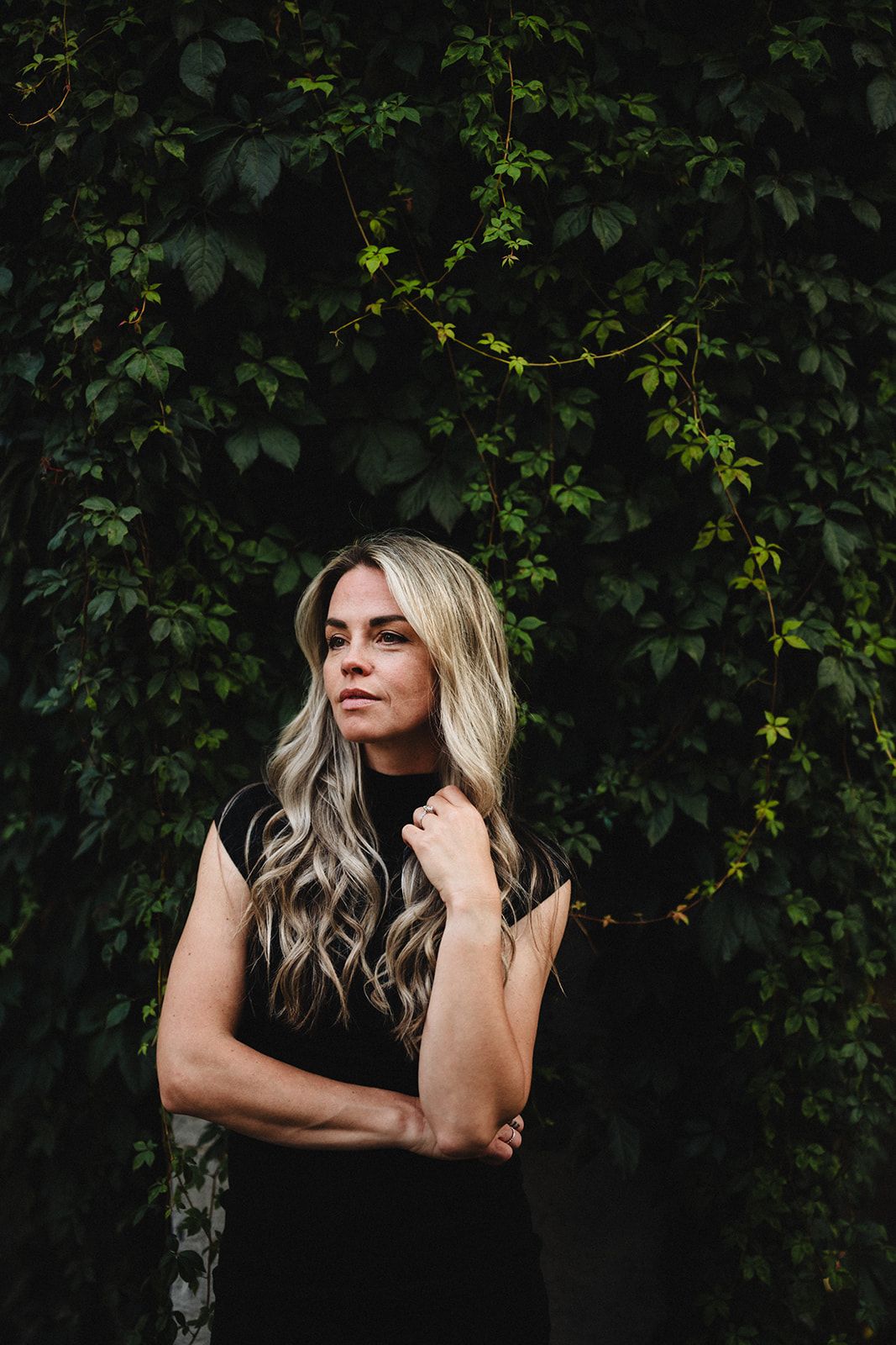A woman with long blonde wavy hair poses against a dark green leafy background in a black sleeveless top.