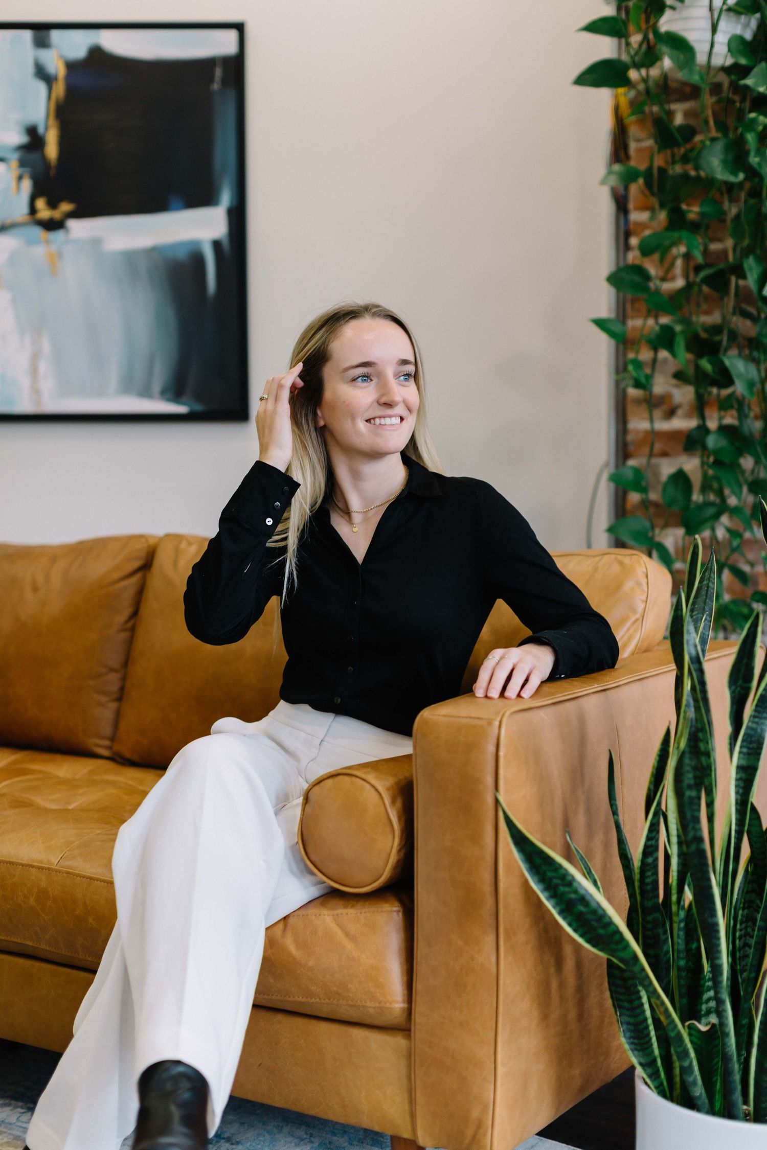 Person in black top relaxes on tan leather couch with indoor plants in background.