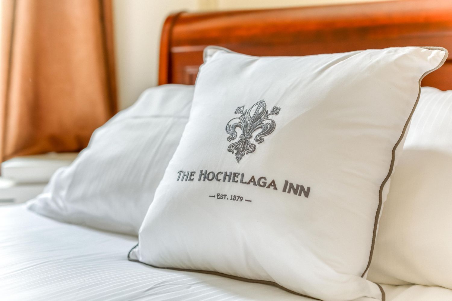White pillow with The Hochelaga Inn logo embroidered on a bed with wooden headboard.