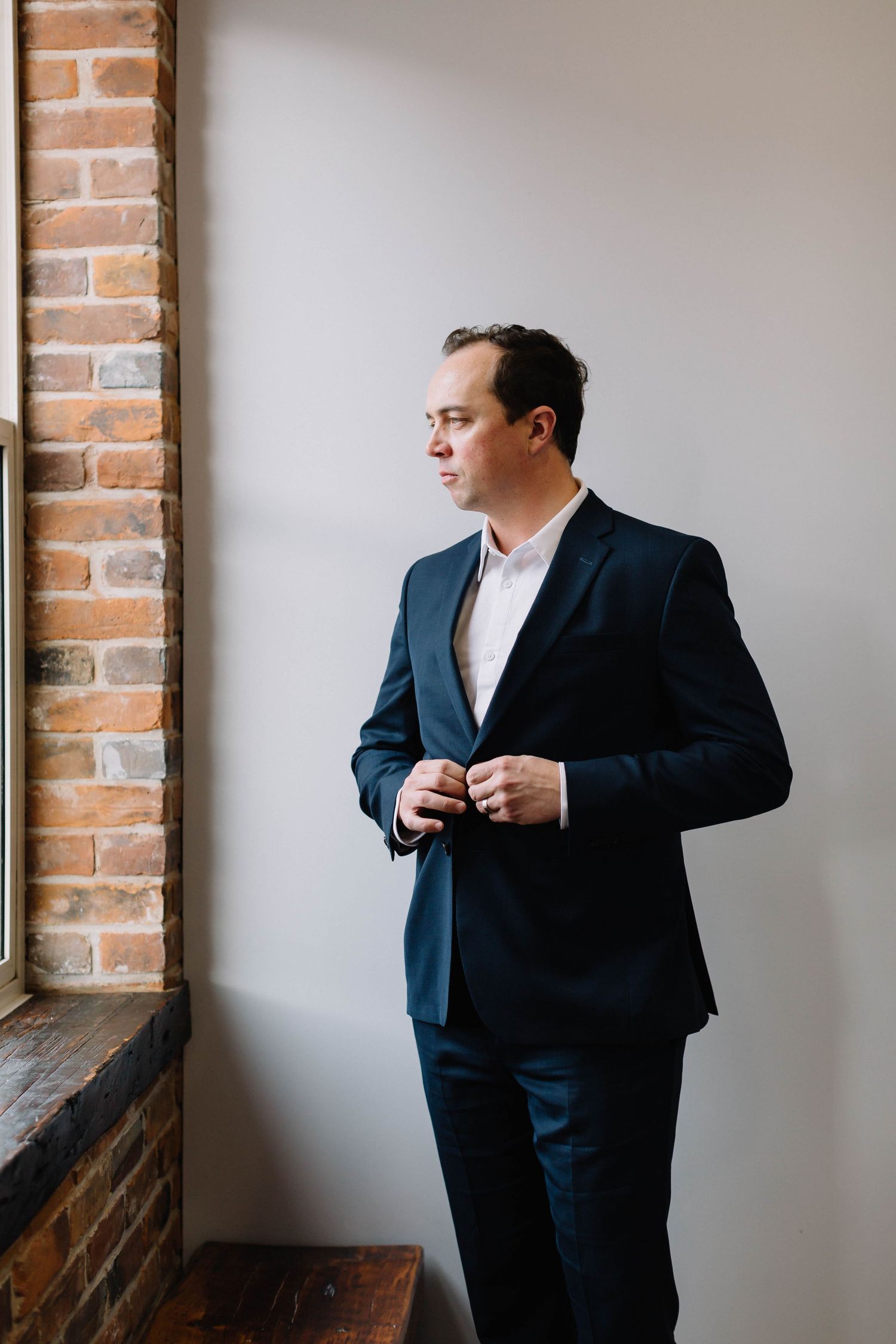 Professional in navy suit adjusts his tie while standing against exposed brick wall in modern office setting.