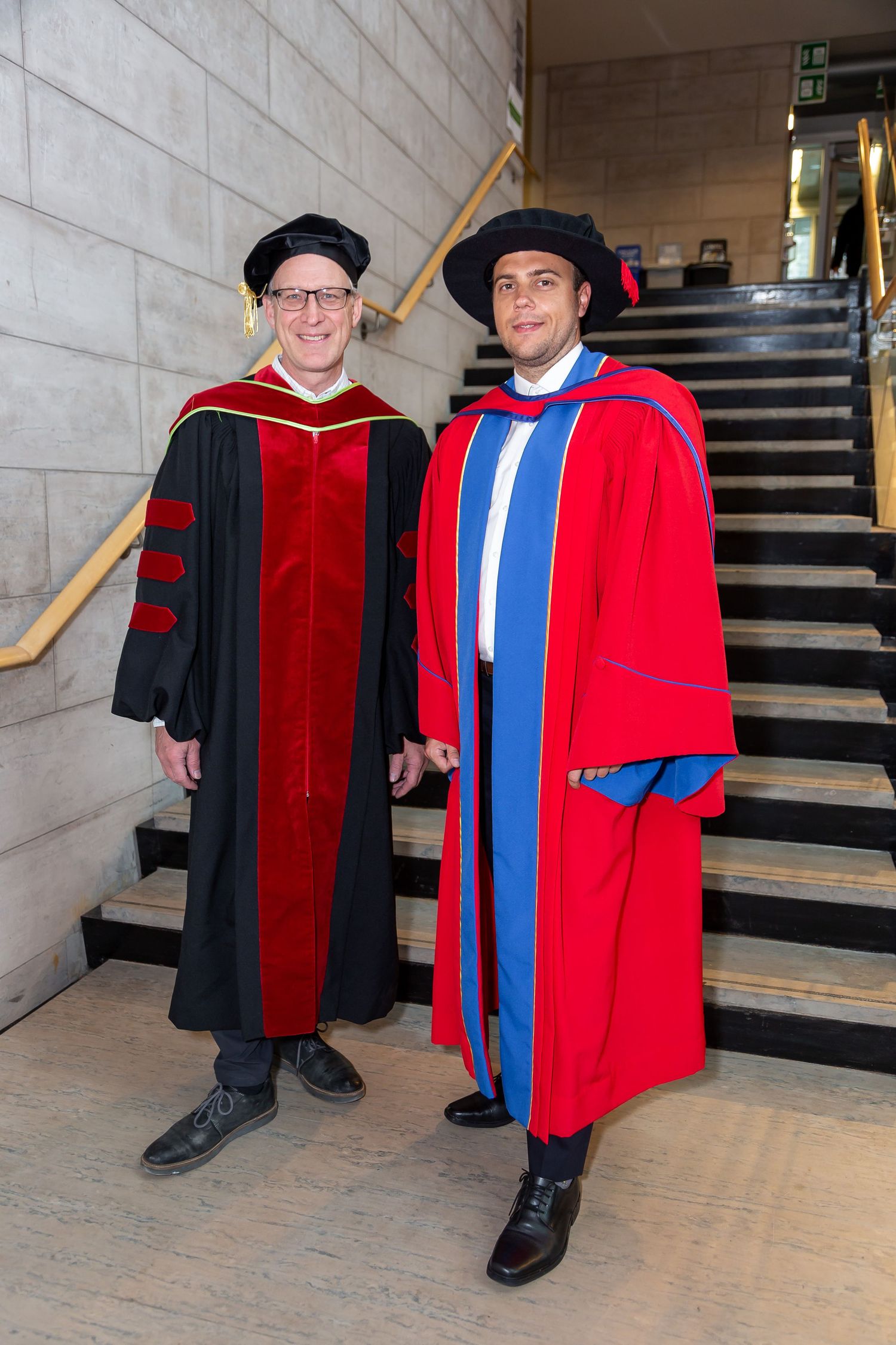 Two graduates in academic regalia standing on stairs in formal convocation attire.