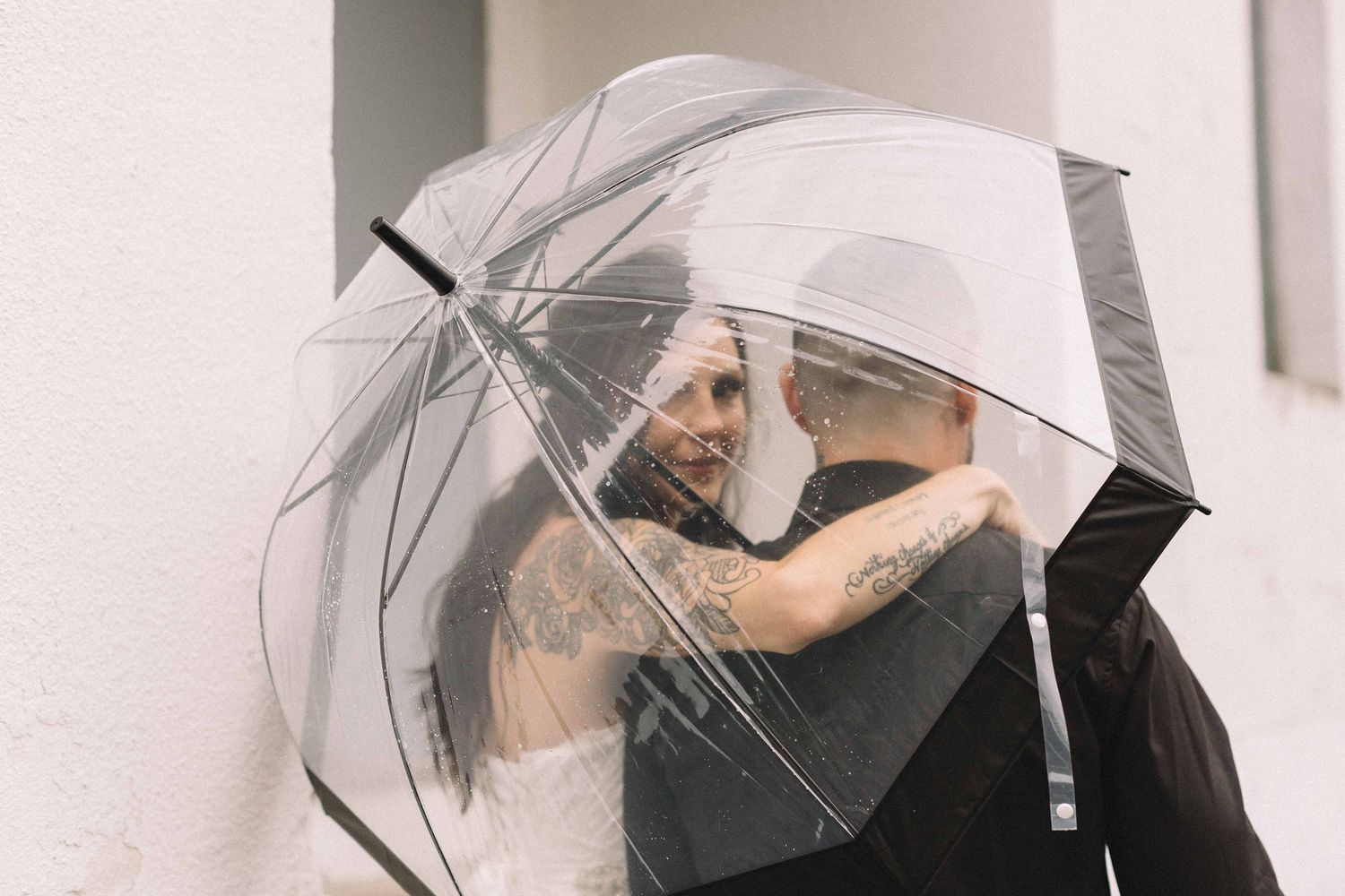 A romantic moment shared under a clear umbrella against a white wall.