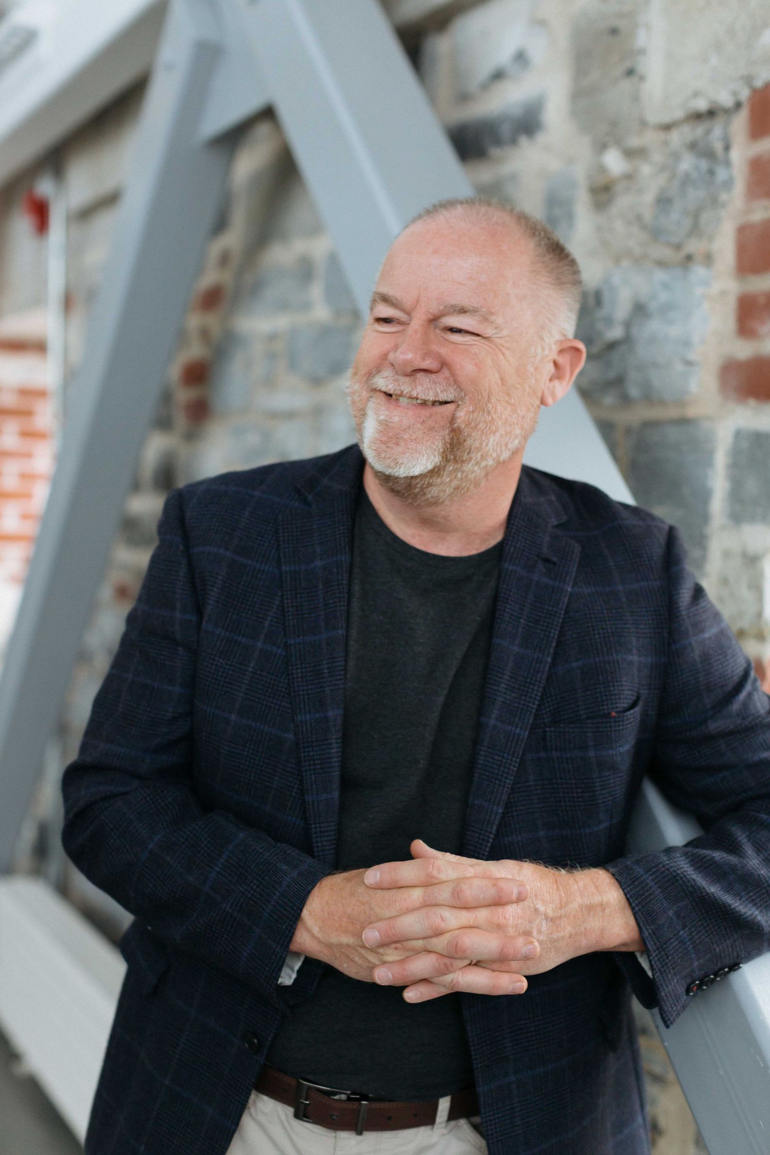 A man in a blazer smiles warmly while standing against a stone and brick wall.