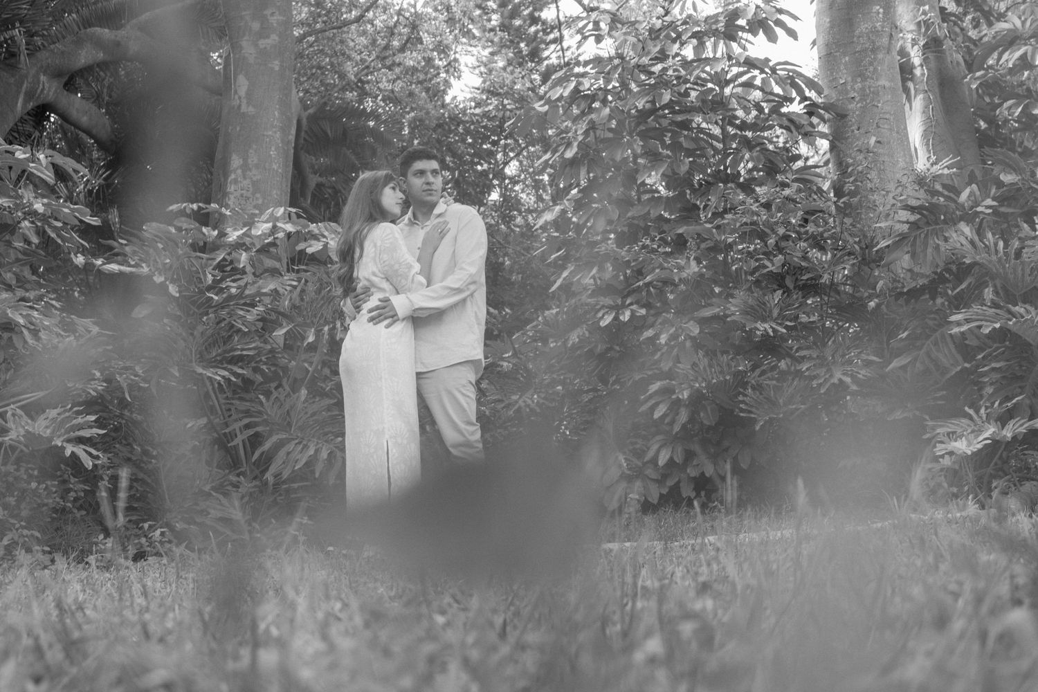 Black and white photo of a couple embracing romantically in a lush garden surrounded by tropical trees and plants.
