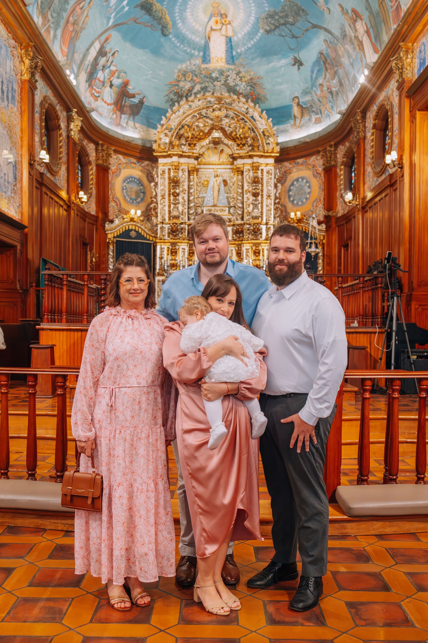 Family posing in ornate church with golden altar, painted dome ceiling, holding baby in christening gown.