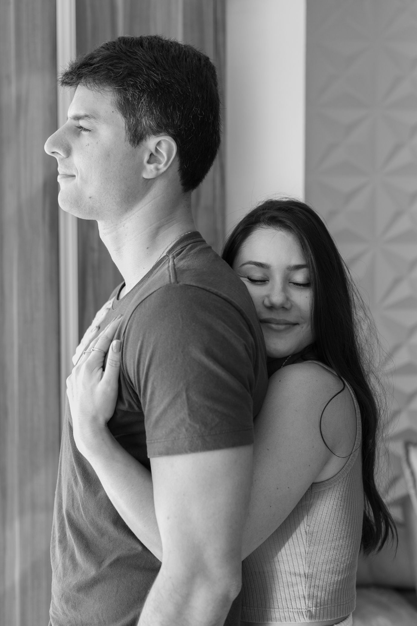 Black and white photo of a couple embracing, woman hugging man from behind with eyes closed indoors.