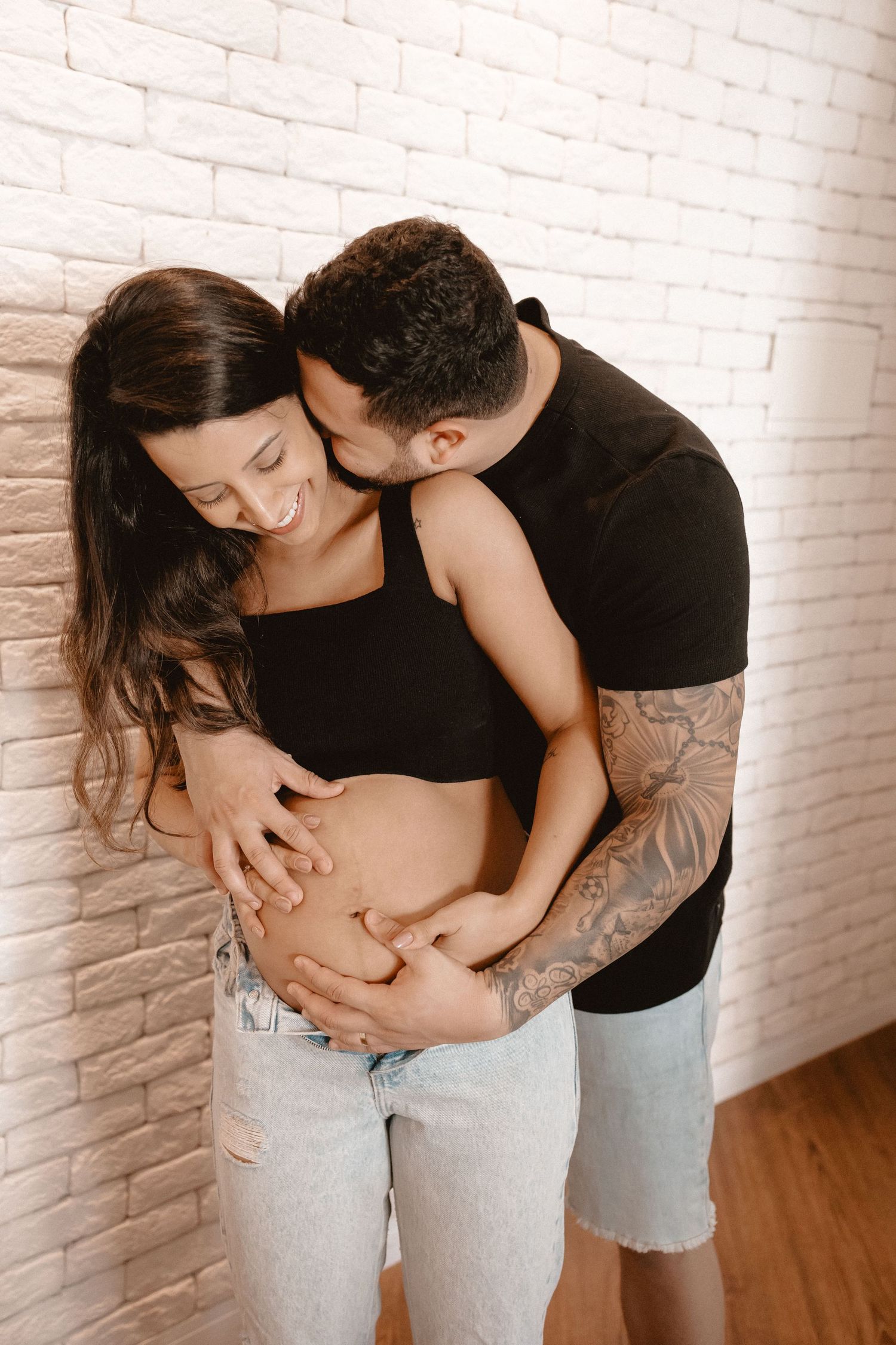 Intimate maternity photo shoot showing couple embracing against white brick wall.