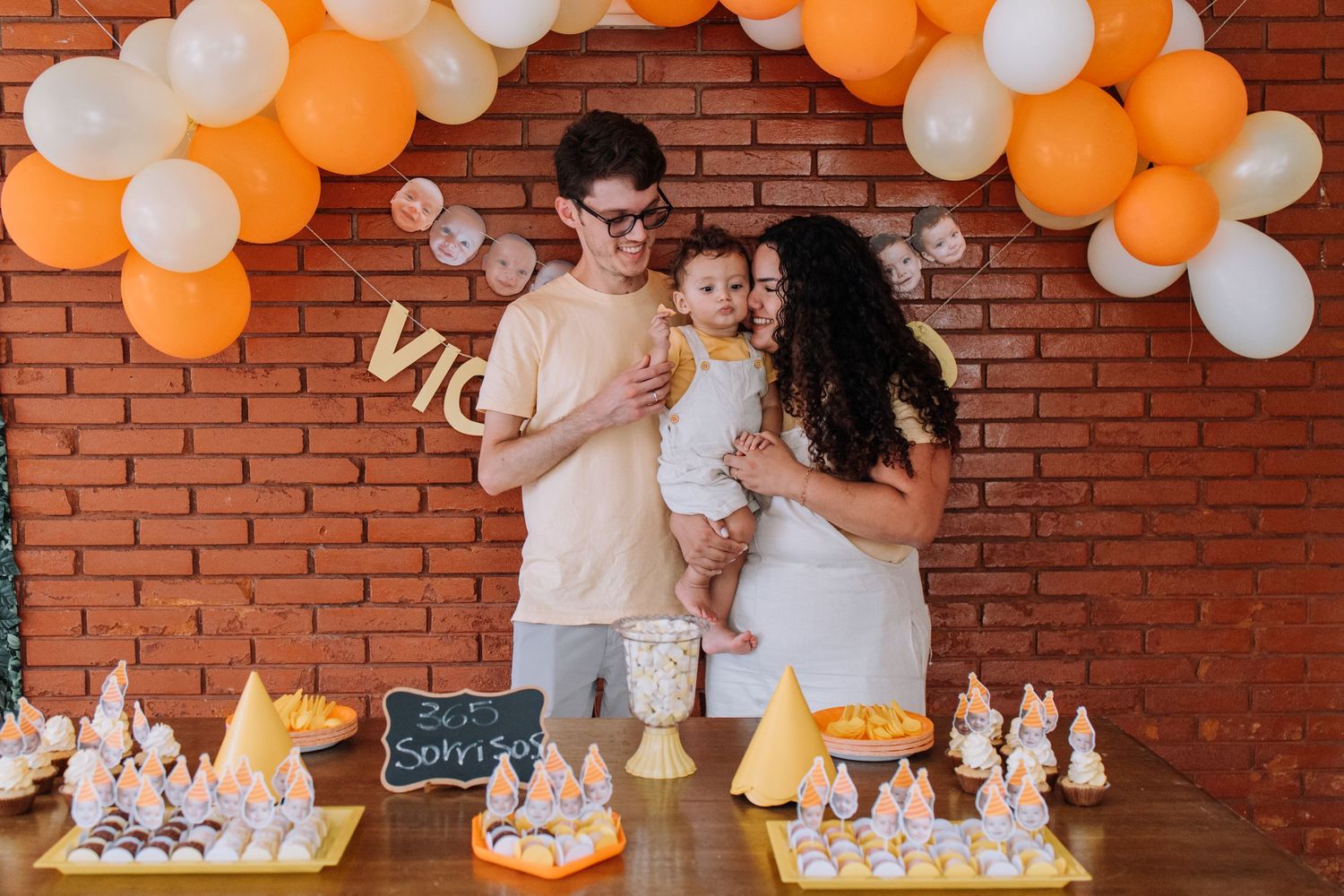 Family celebrating at birthday party with orange and white balloon arch and festive decorations.