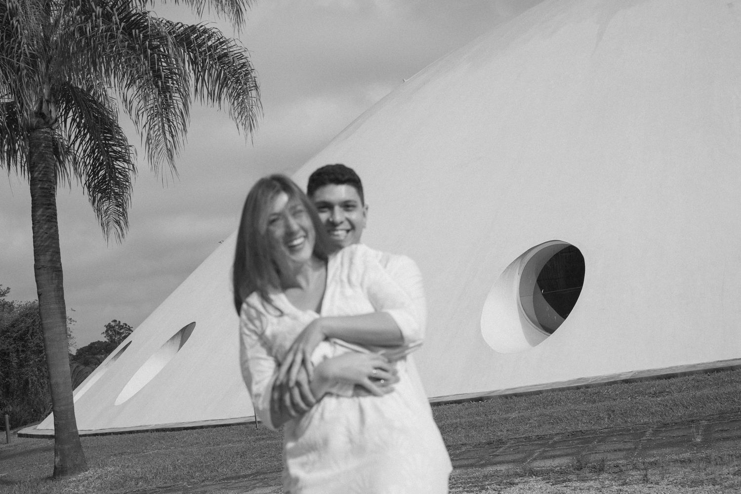 Couple embracing and smiling in white outfits outdoors near a palm tree and modern white building.