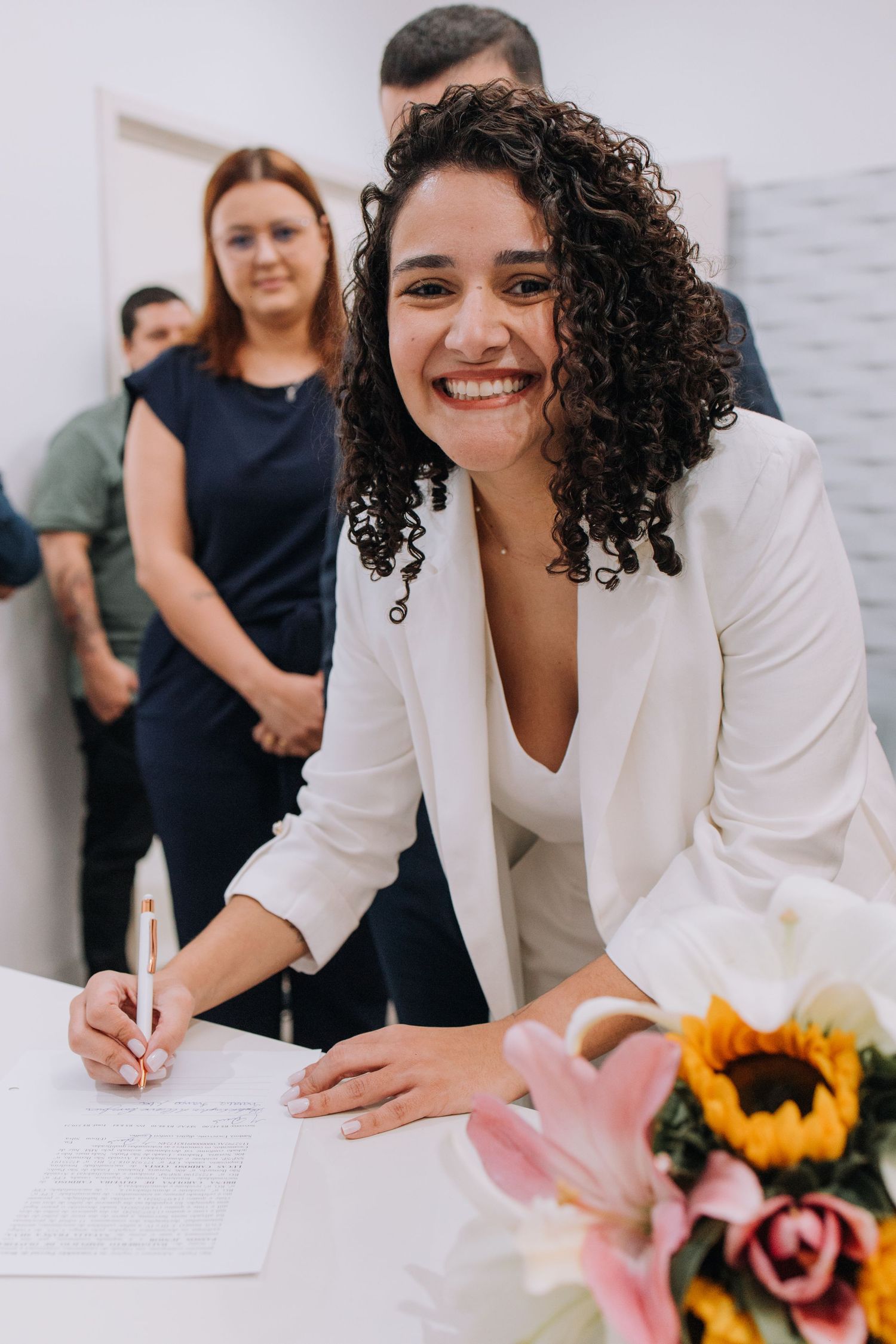 Three sequential images show someone signing documents at a desk with colorful flowers in the foreground.
