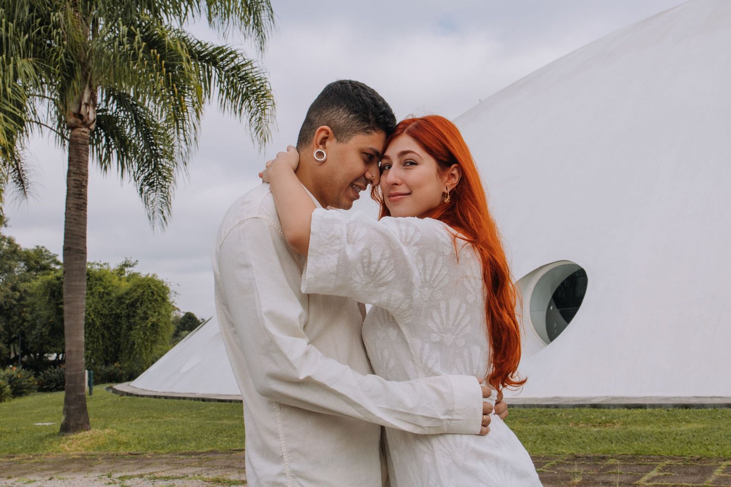 Couple in white outfits embracing near palm trees and a white dome building on a cloudy day.