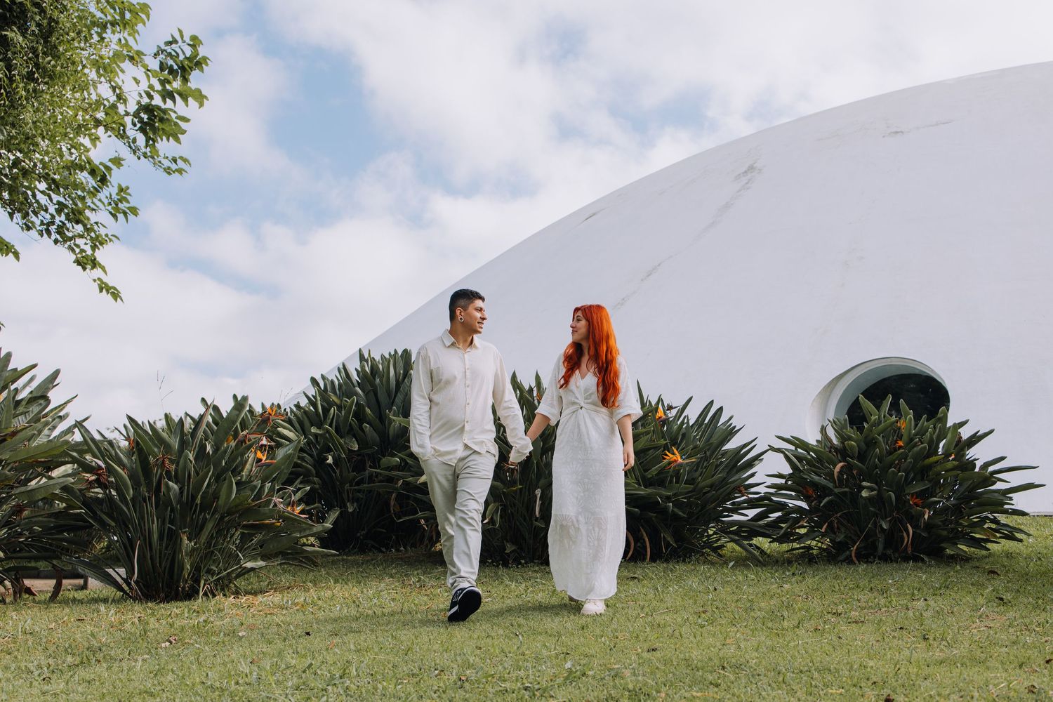 Couple in white wedding attire walking on green lawn beside a white dome building with tropical plants.
