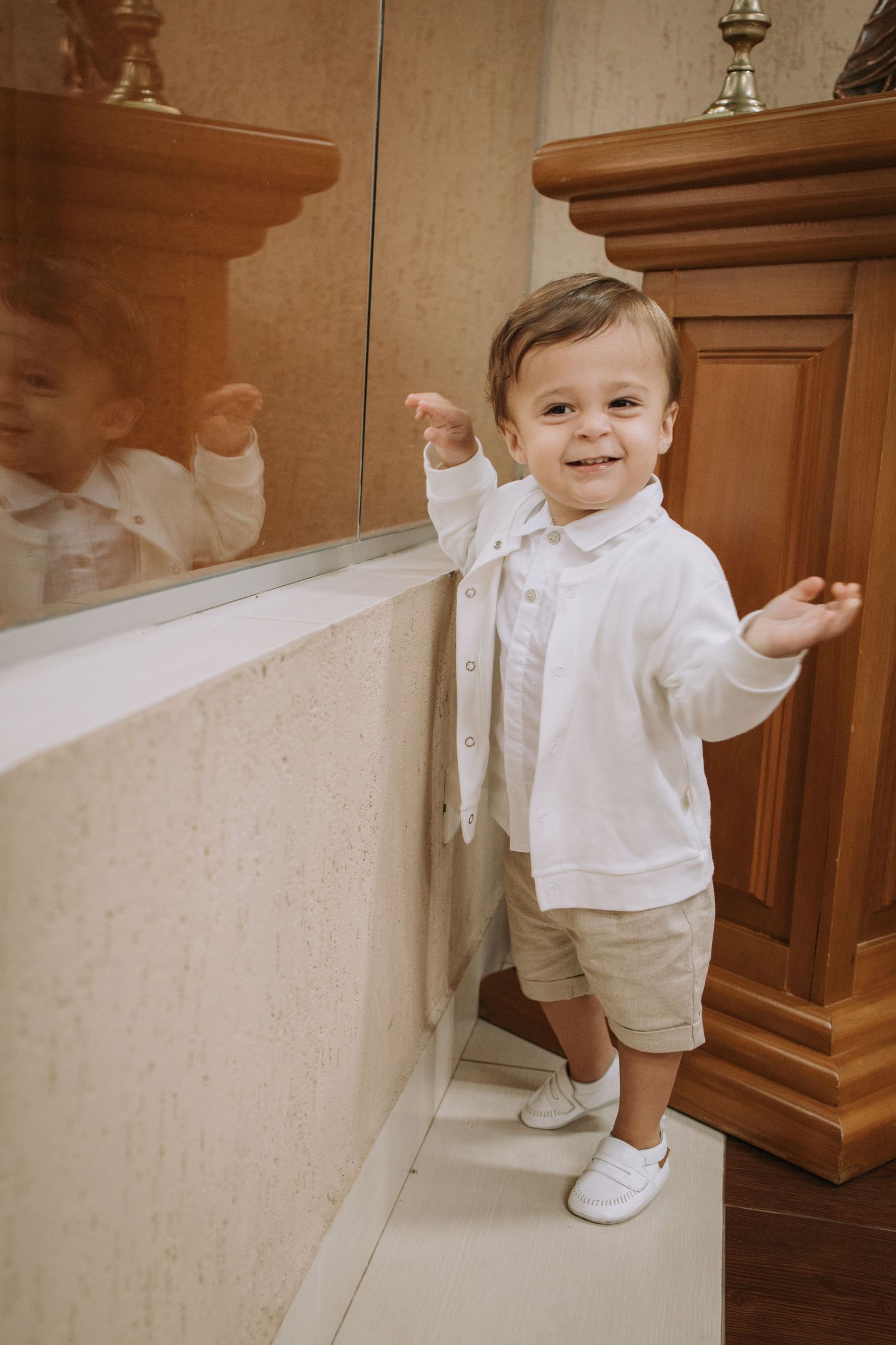 A toddler in white clothing walks along a wall in an indoor setting.