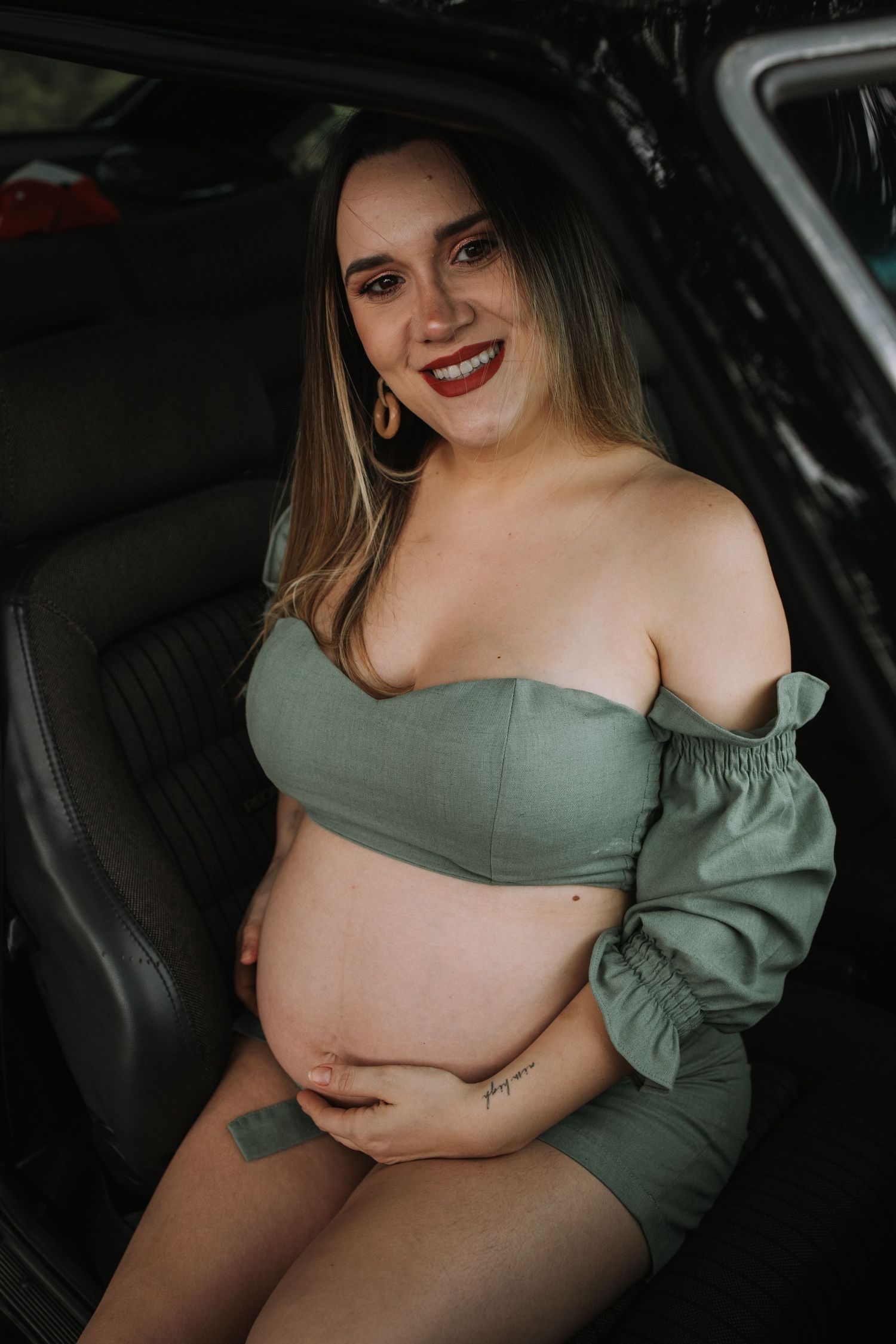 Maternity portraits taken inside car showing woman in sage green off-shoulder dress.