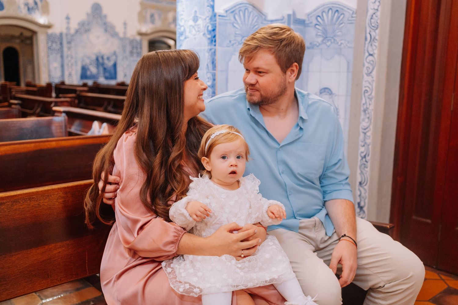 Family with baby girl in white dress sitting in church pew during baptism ceremony.