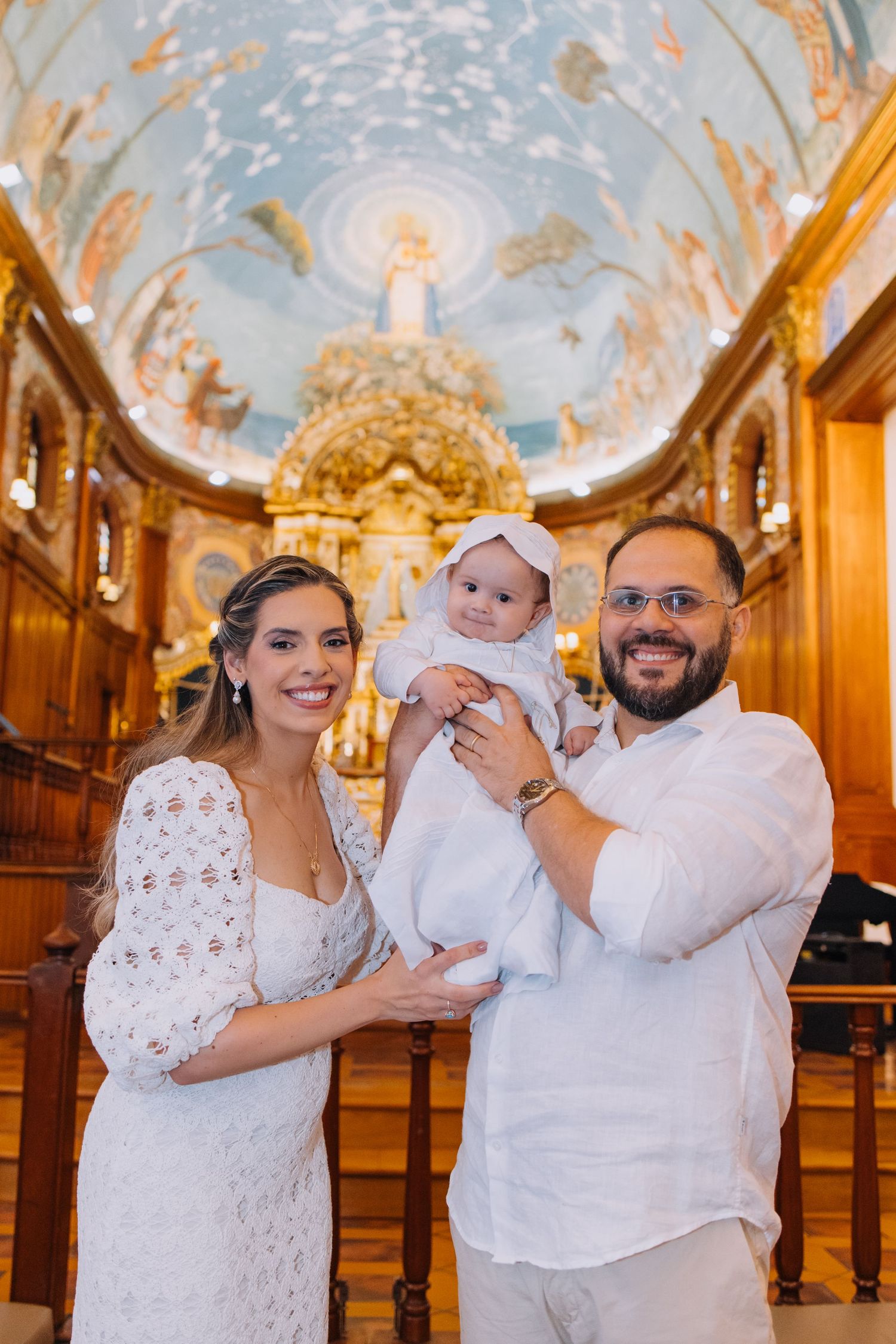 Family posing with baby in white baptism gown inside ornate church with painted ceiling and golden altar.