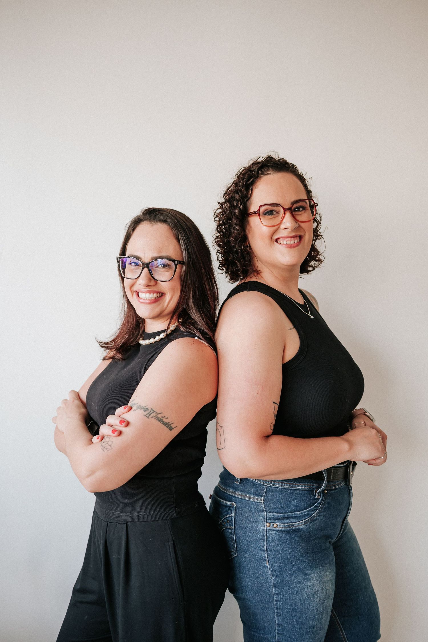 Two women in glasses pose back-to-back with crossed arms against a white wall, wearing black tops and casual attire.