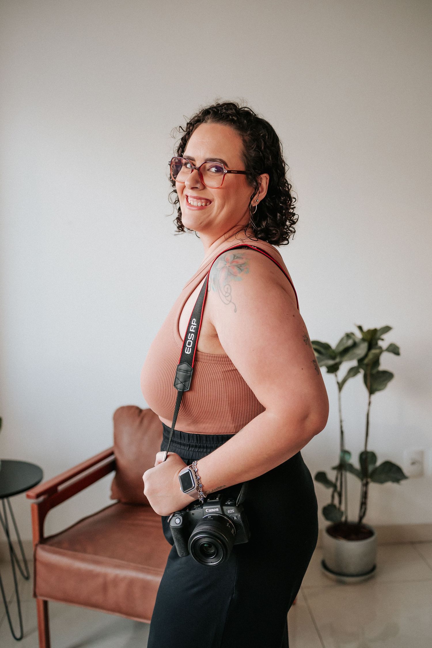 A person in a black dress poses against a white wall with a brown leather chair and houseplant visible in the background.