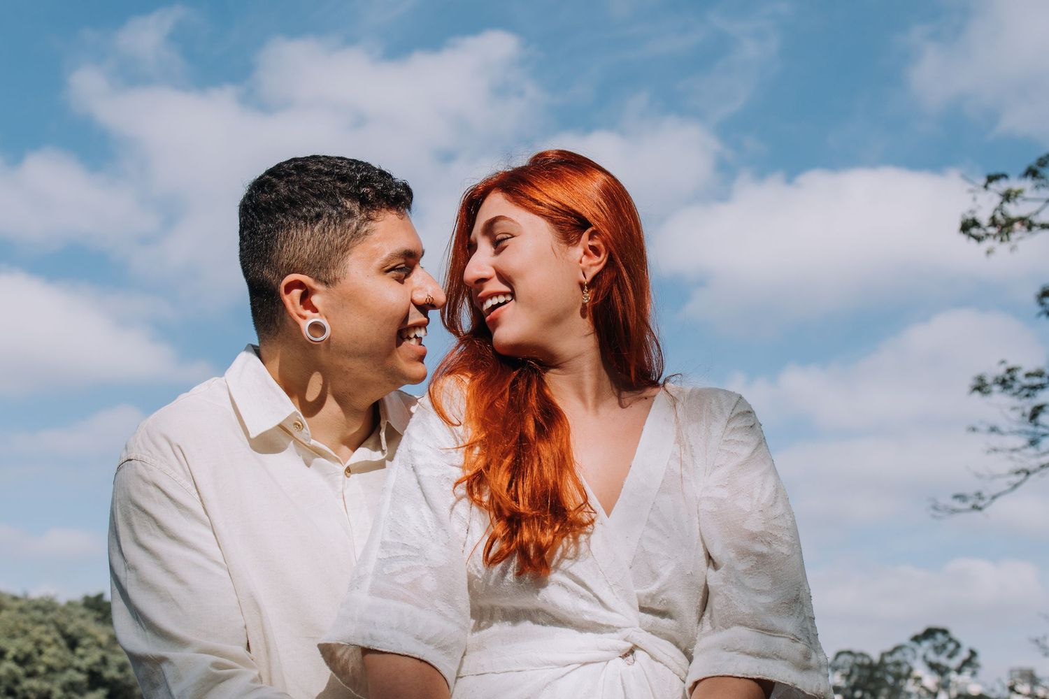 Two women smiling at each other outdoors, one with red hair, wearing white outfits under a blue sky.