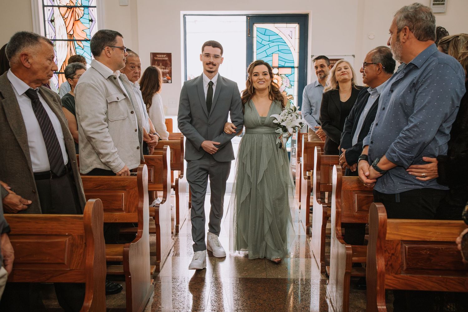 A couple stands in the center aisle of a small chapel with wooden pews and guests looking on during a ceremony.