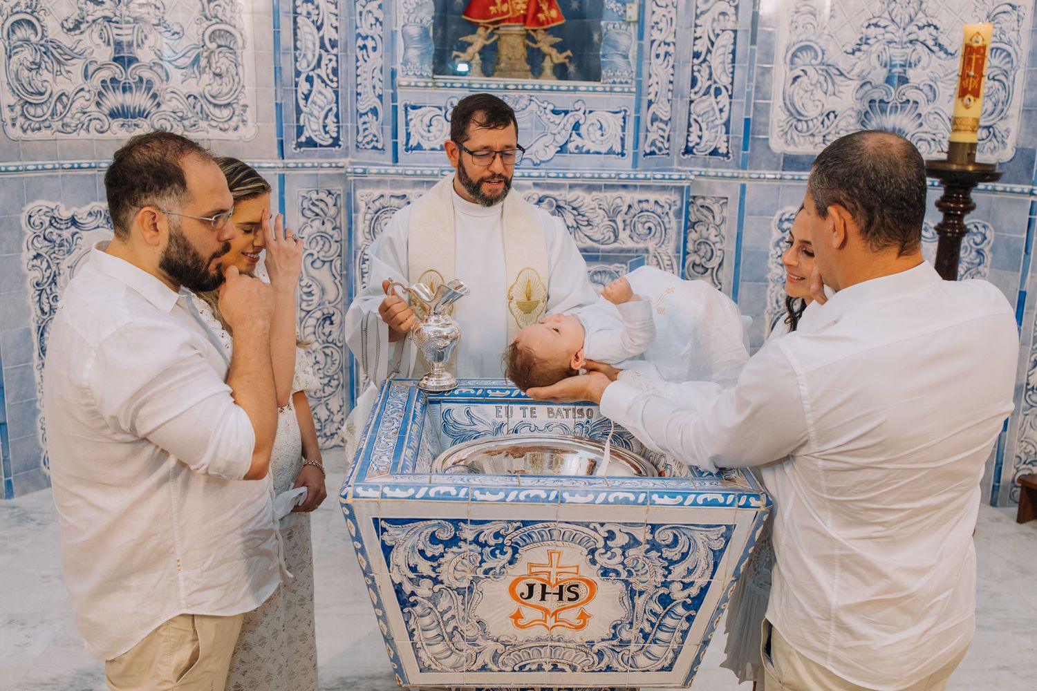 Catholic baptism ceremony with priest and family gathered around ornate JHS font in blue tiled church.