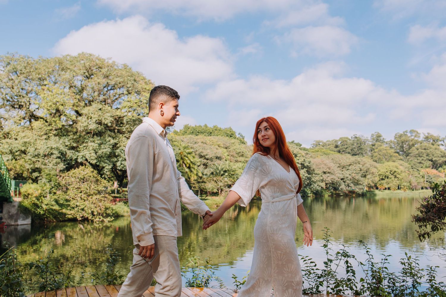 Couple holding hands by a serene lake surrounded by lush green trees under a partly cloudy sky.