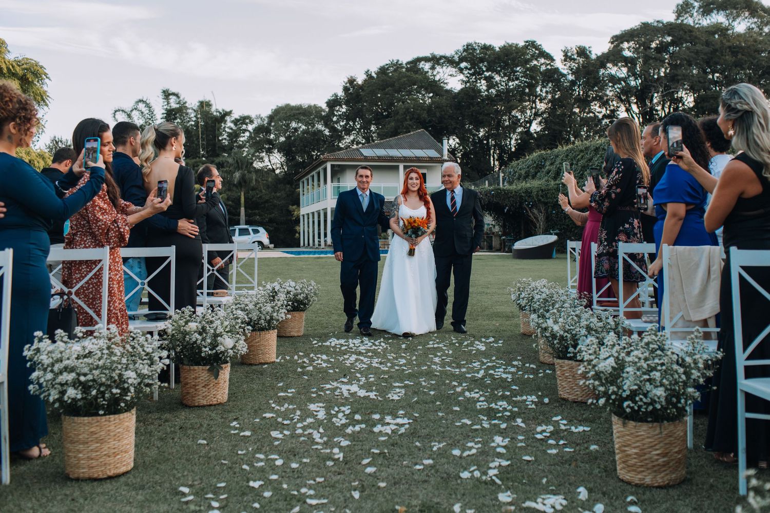 Couple walks down outdoor aisle lined with rustic flower barrels as wedding guests capture the moment on phones.