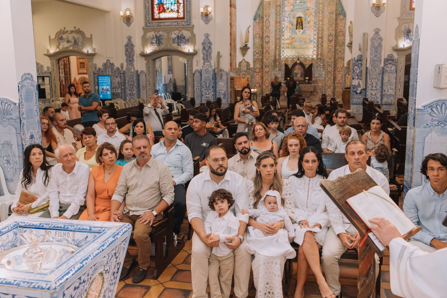 Families gathered in an ornate church with blue tile walls for a baptism ceremony, dressed in white attire.