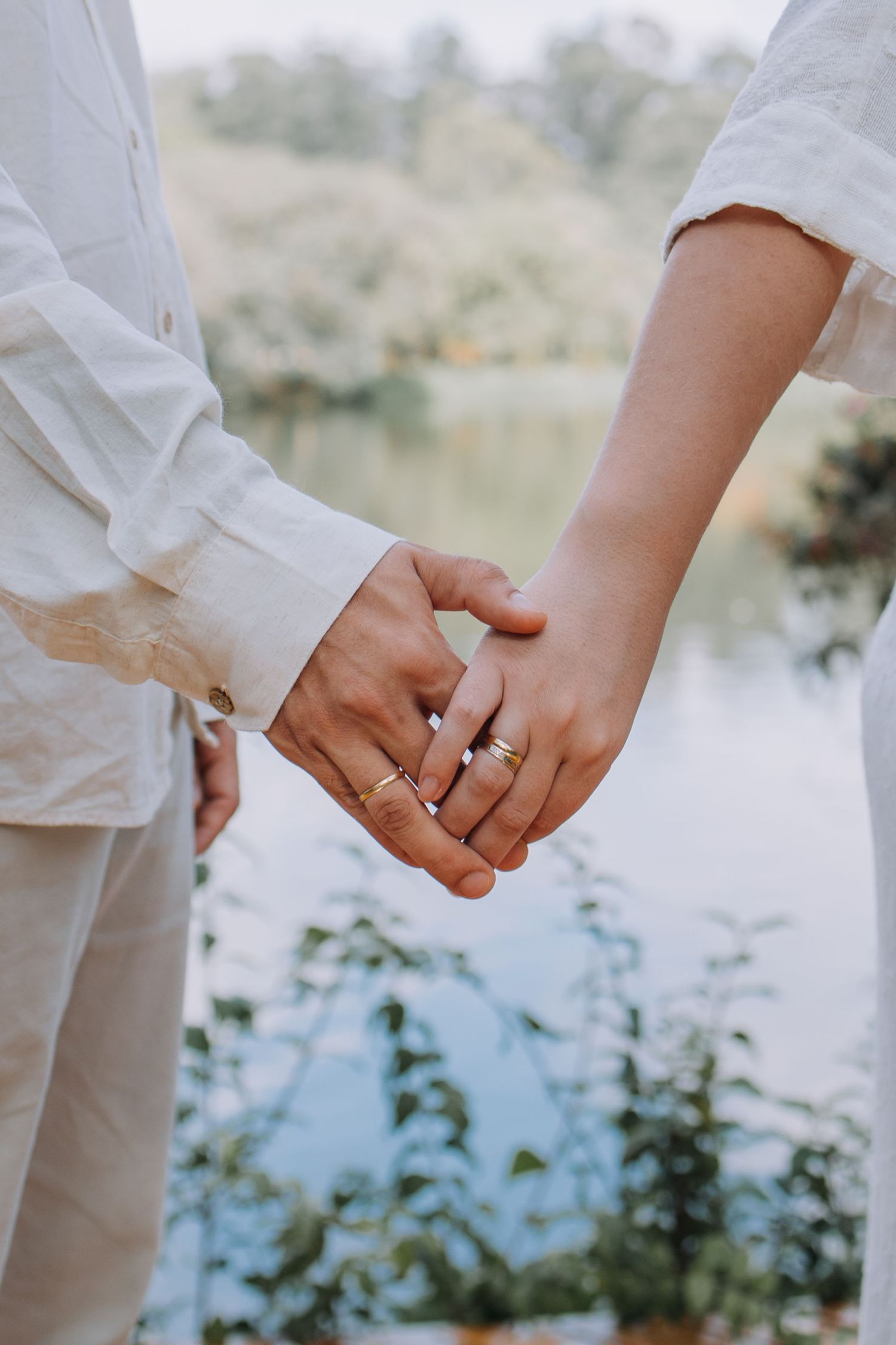 Couple holding hands near a lake, wearing white outfits and gold rings, romantic outdoor setting.