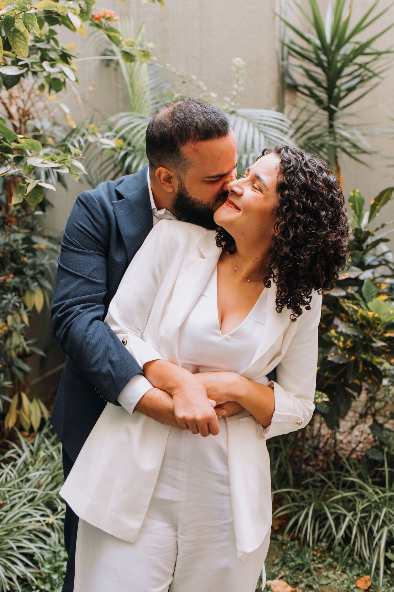 A couple in coordinated white outfits share intimate moments in a garden setting with tropical plants.
