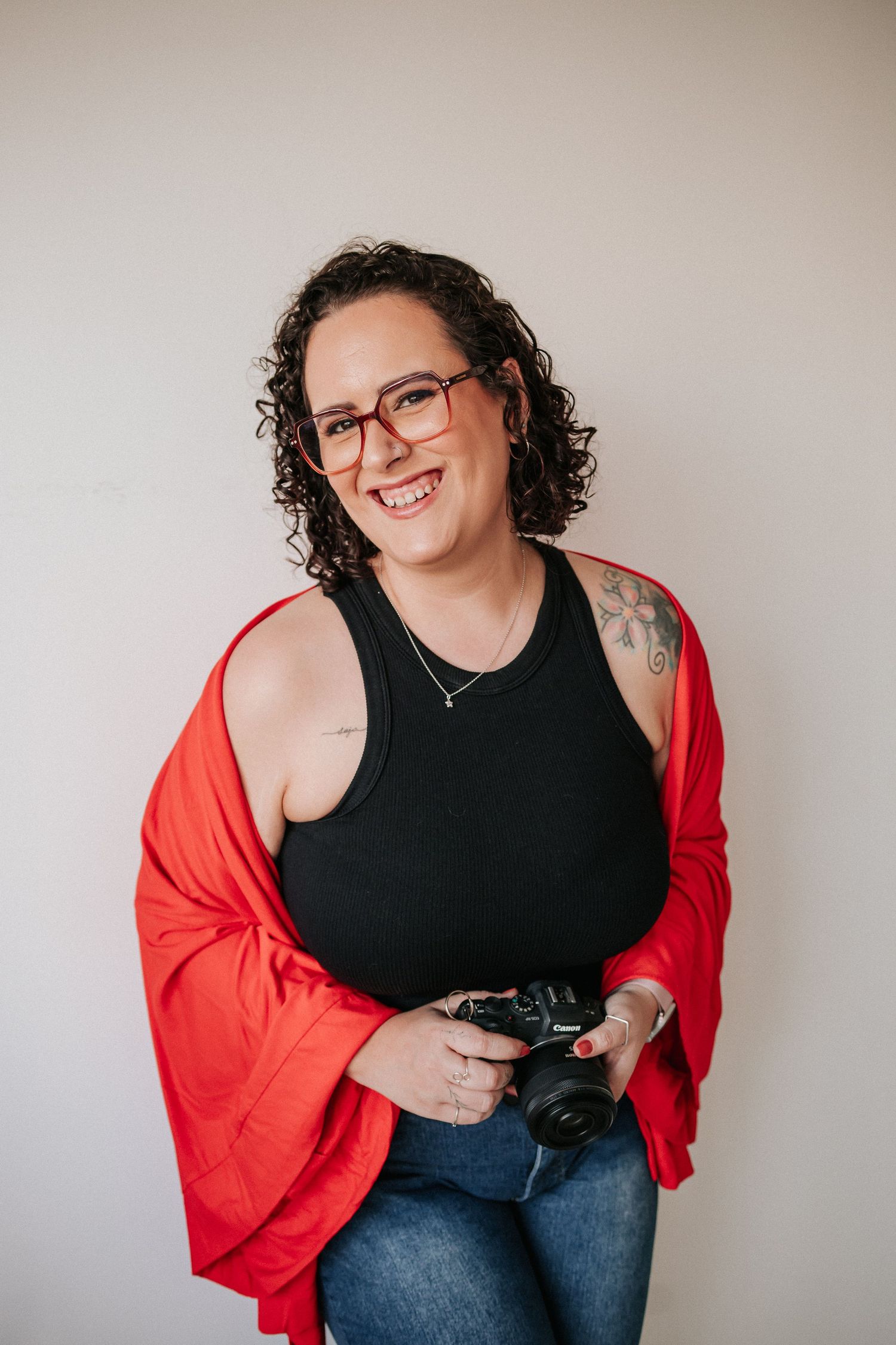 A photographer wearing a red cardigan and black top holds a camera while sitting against a white wall.