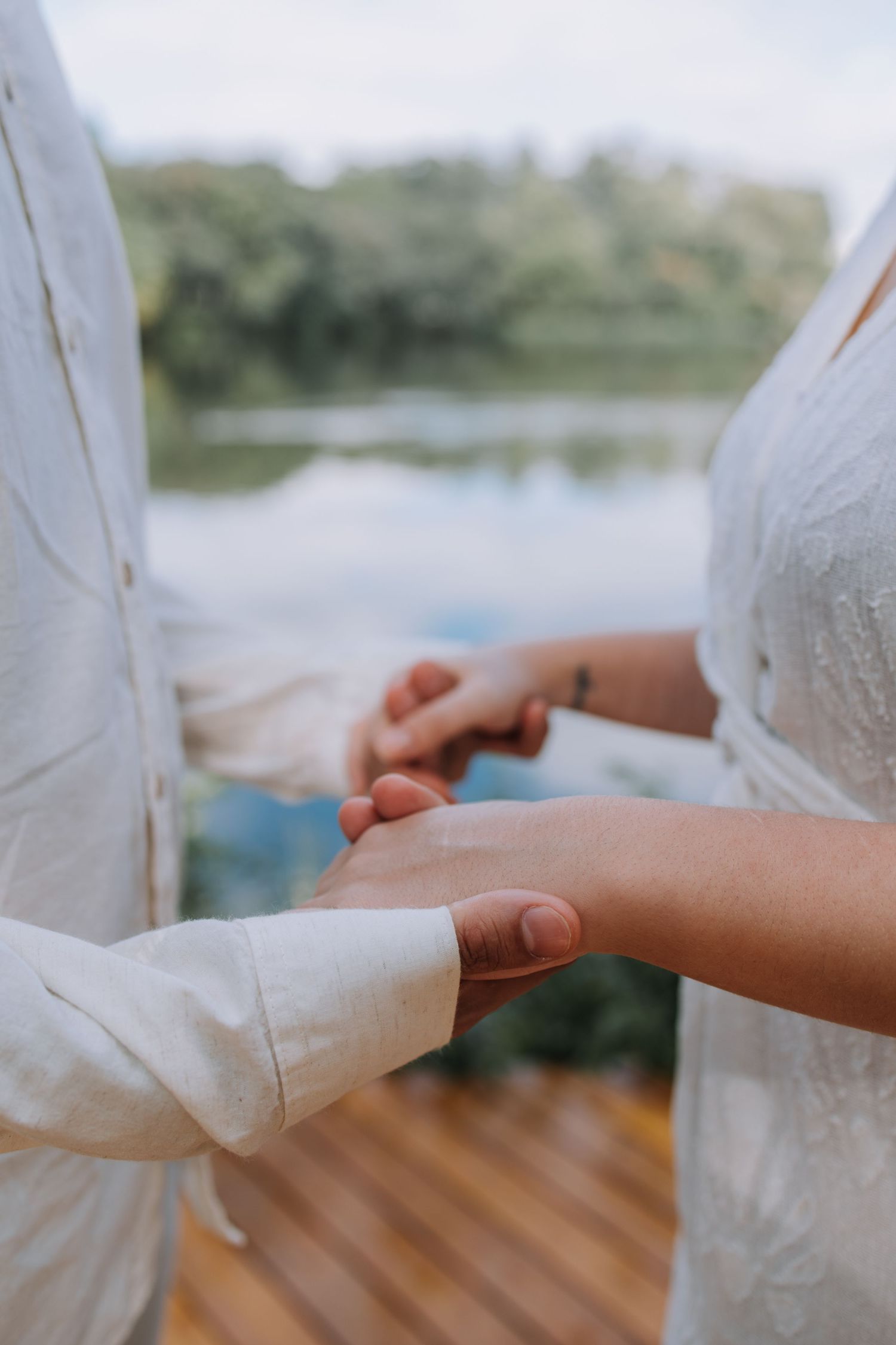Couple exchanging rings on a wooden deck by a lake, both dressed in white during an outdoor wedding ceremony.