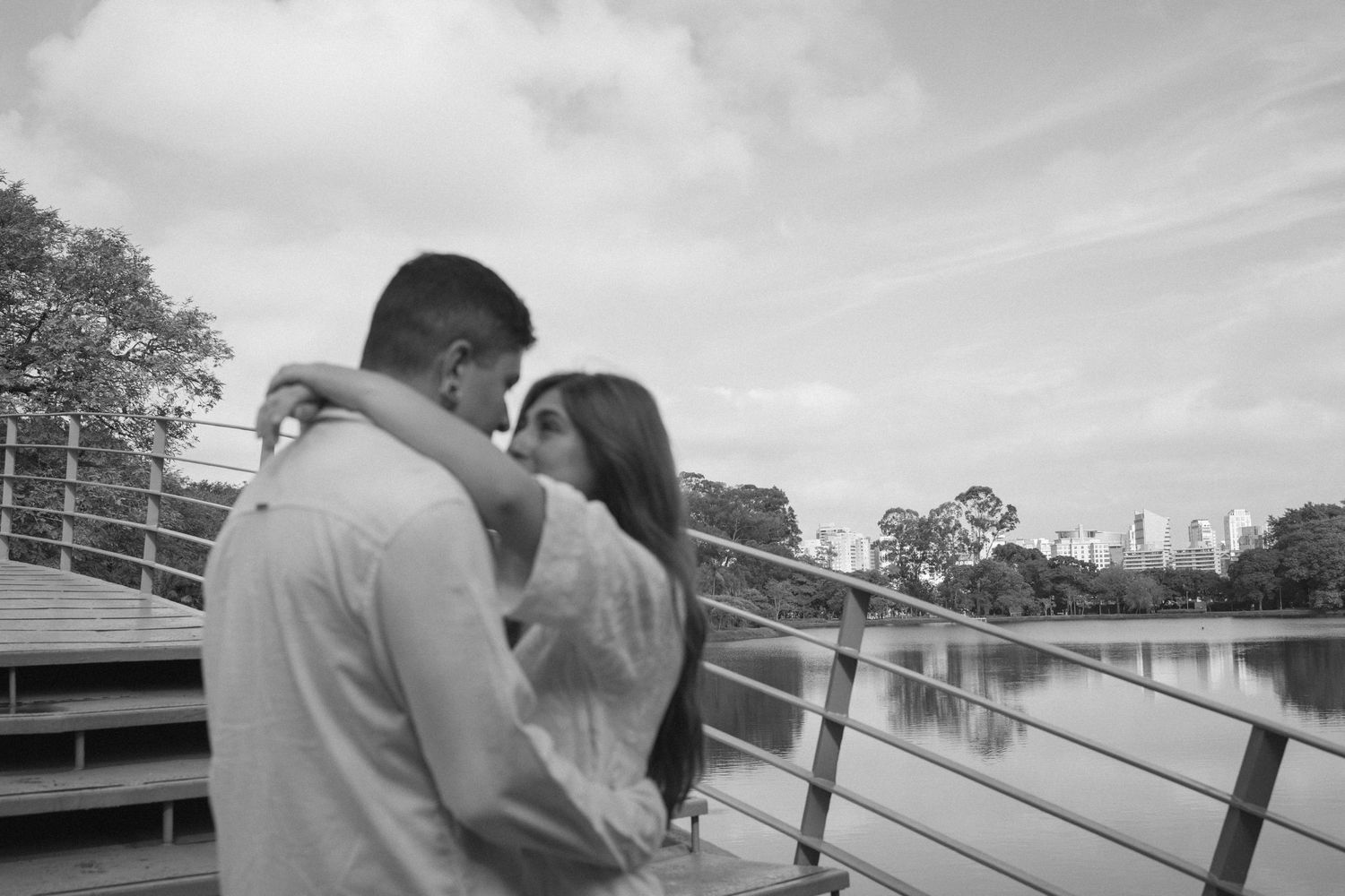 Black and white photo of a couple sharing a romantic moment on a bridge overlooking a serene lake and city skyline.