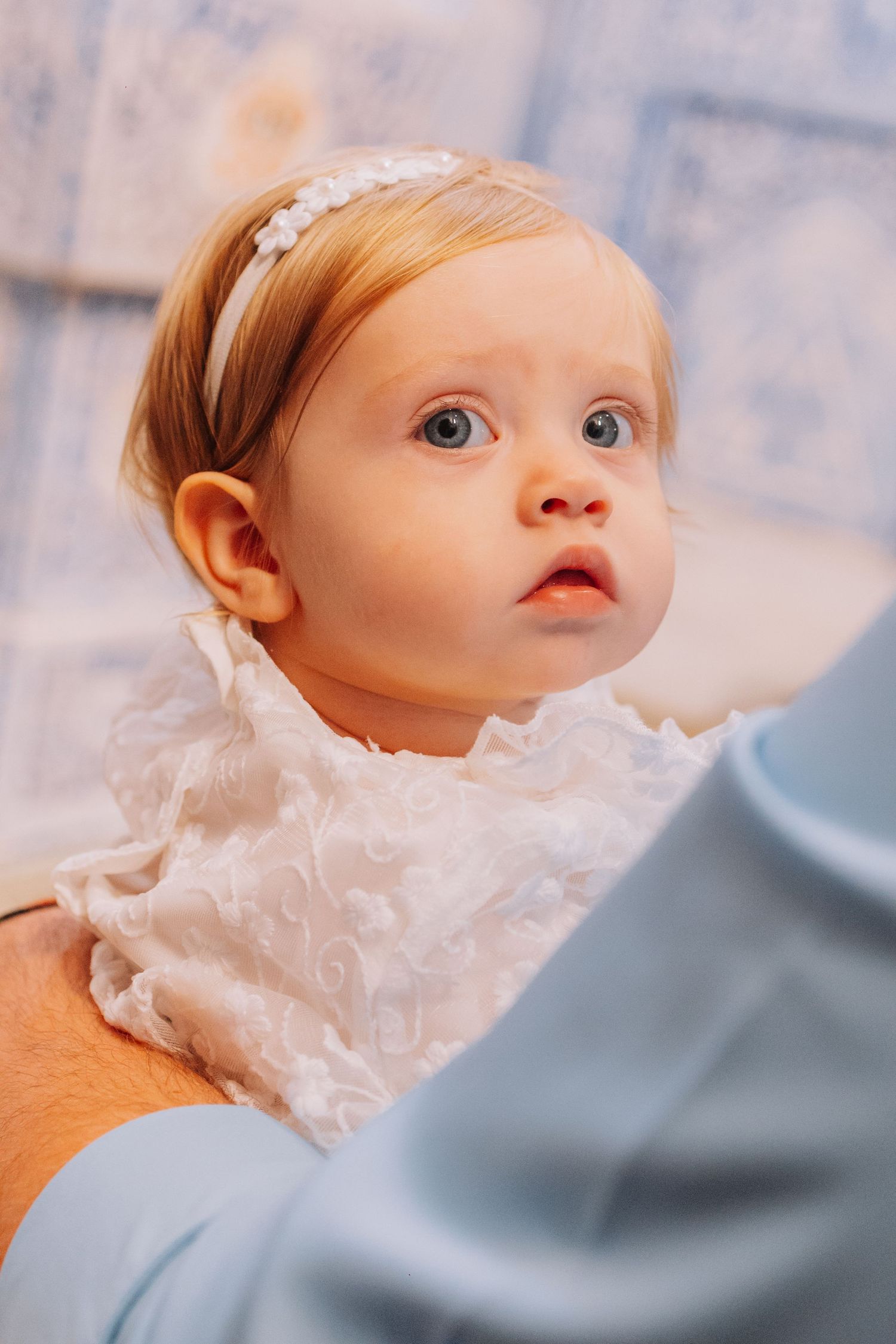Adorable baby girl with blue eyes wearing a white lace outfit and floral headband, looking up with curiosity.