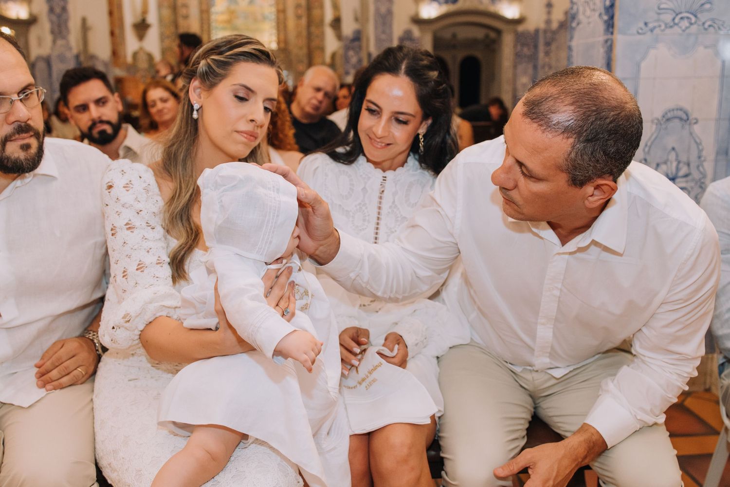Family gathered for a baby baptism ceremony in a church, dressed in white outfits, tenderly touching the infant.