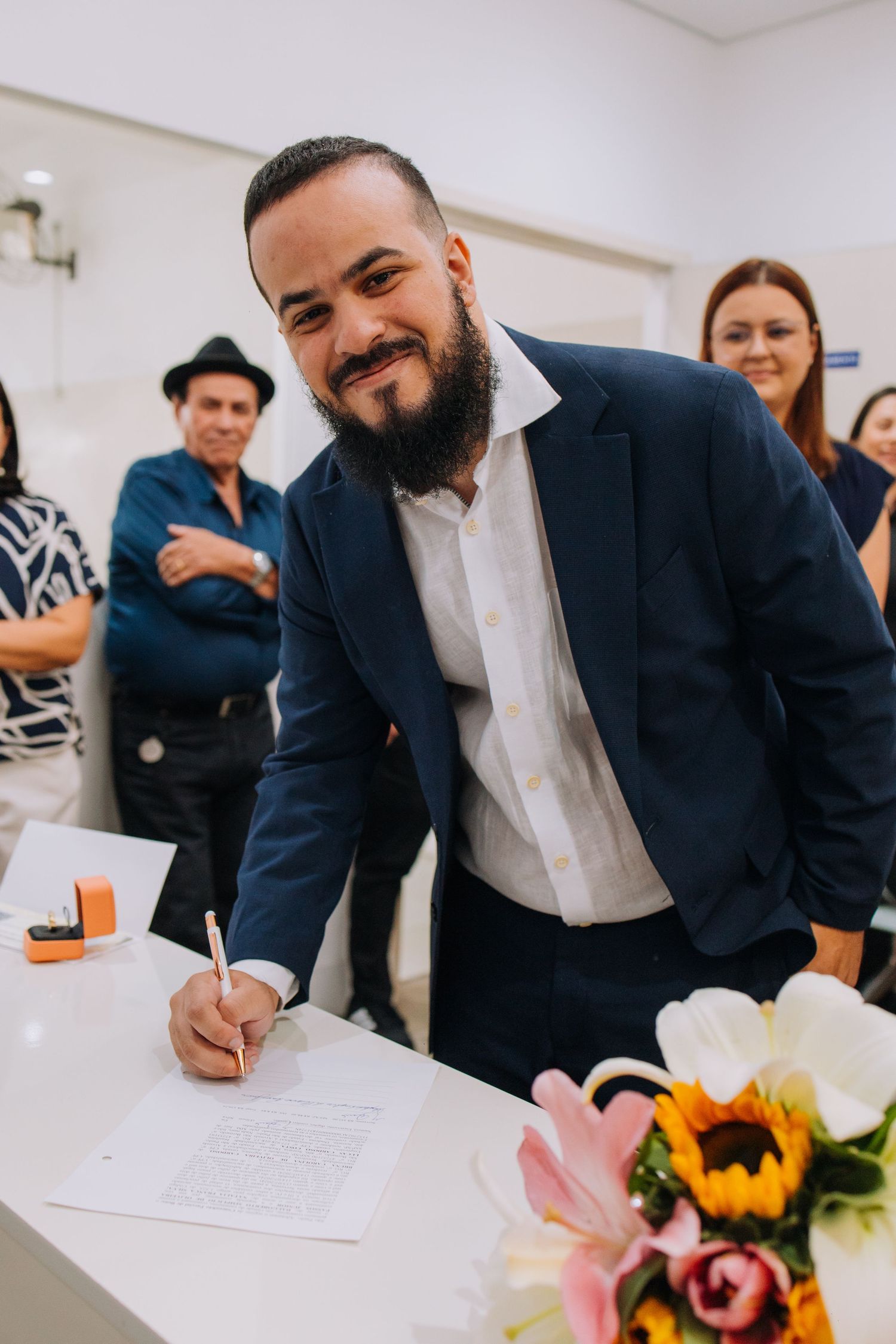 People in navy suits sign documents at a table decorated with colorful floral arrangements during a formal event.