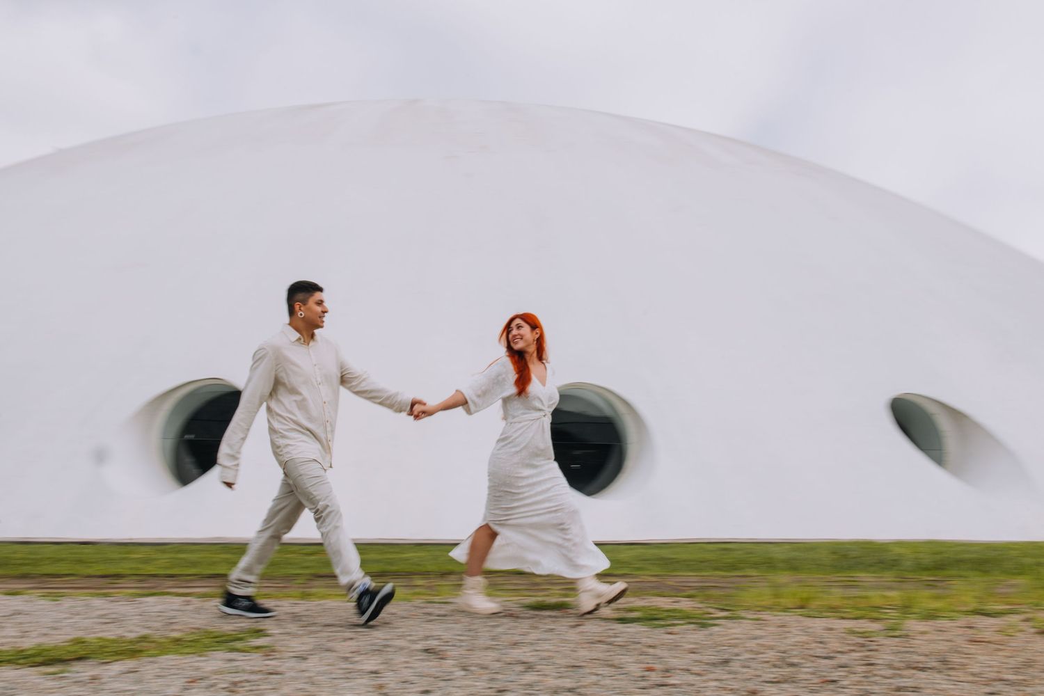 A joyful couple in white wedding attire runs hand-in-hand past a modern white domed building outdoors.