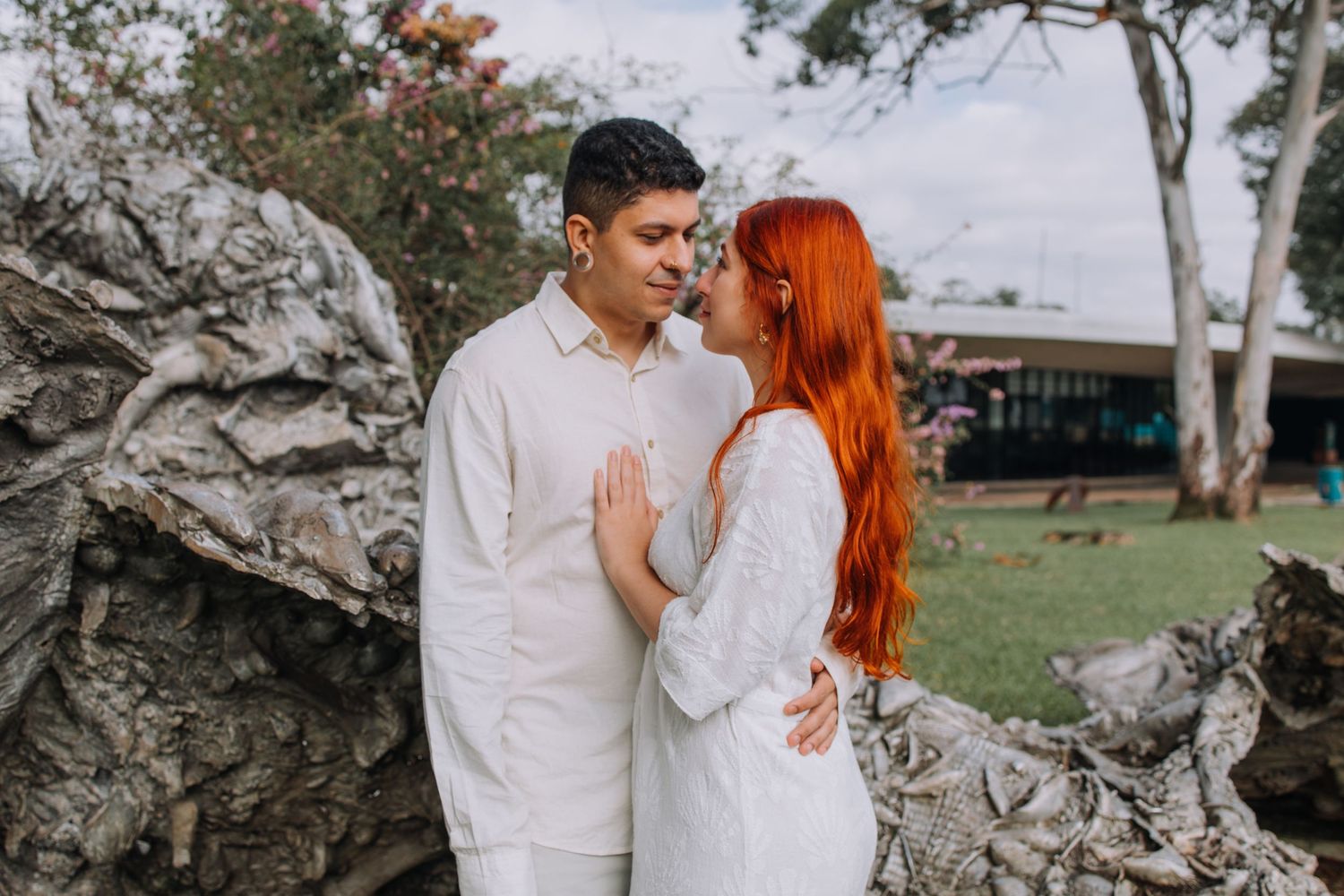 Couple in white outfits sharing a romantic moment near driftwood, woman with vibrant red hair leaning in for a kiss.