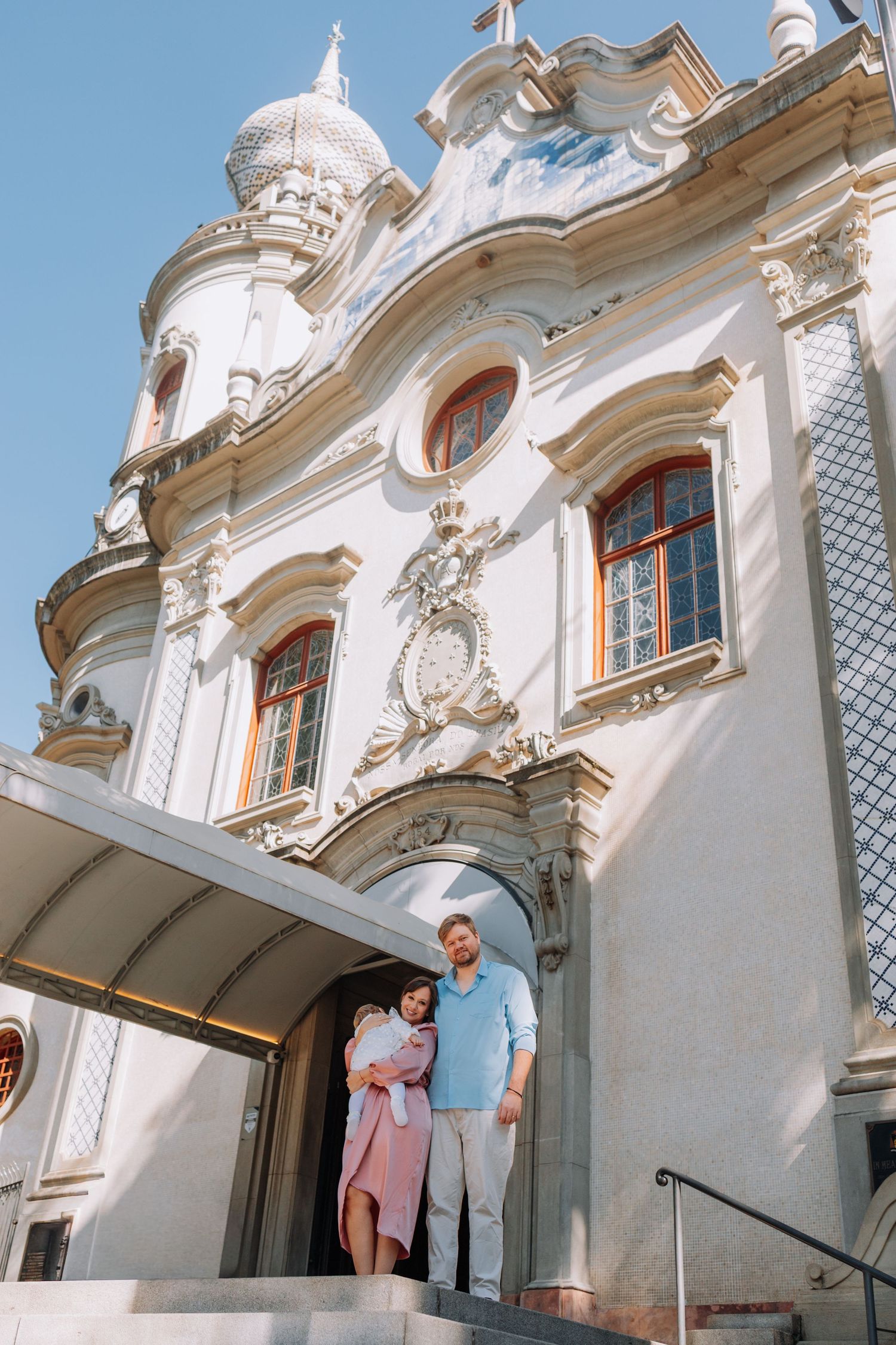 Couple standing on steps of ornate Baroque church facade with white stucco decorations and blue sky.