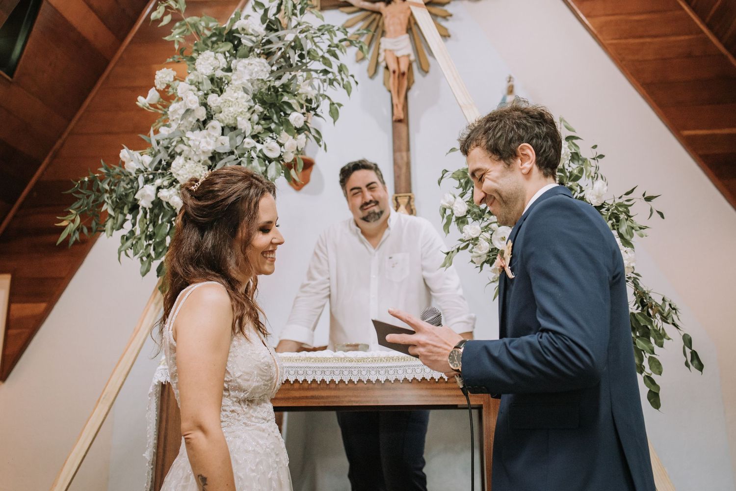 Couple exchanges vows during intimate indoor wedding ceremony with wooden beams and floral decorations.