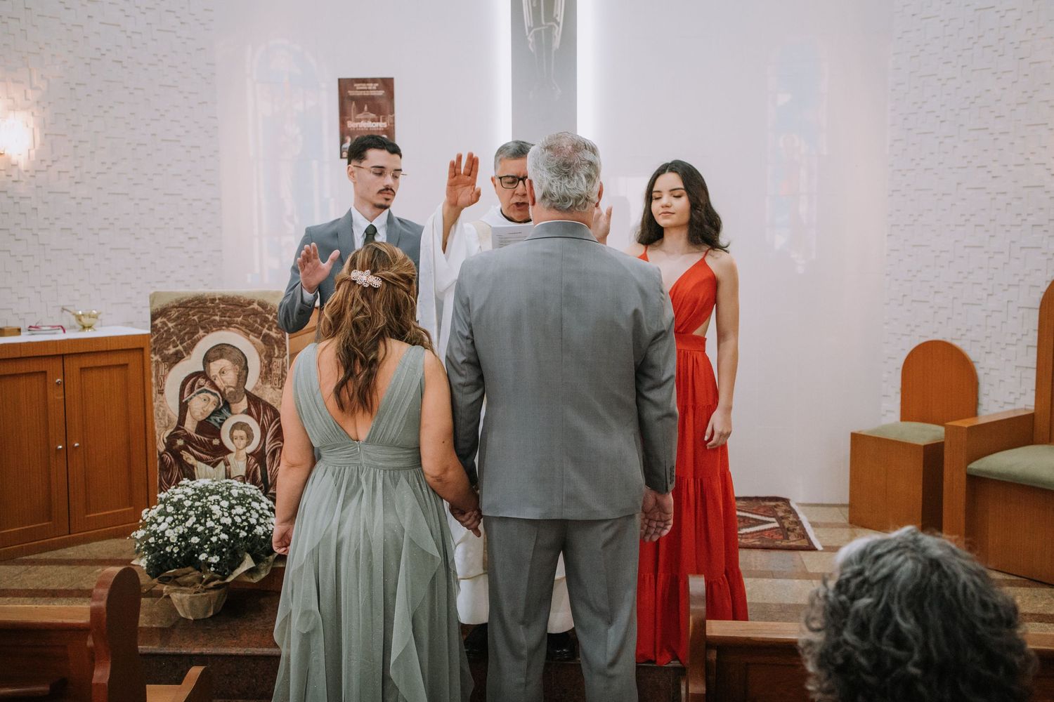 A wedding ceremony takes place in a church with attendees in formal attire including grey and red dresses.