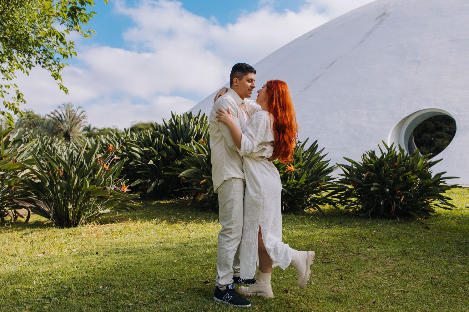 Couple embracing outdoors near white building surrounded by tropical greenery under blue sky.