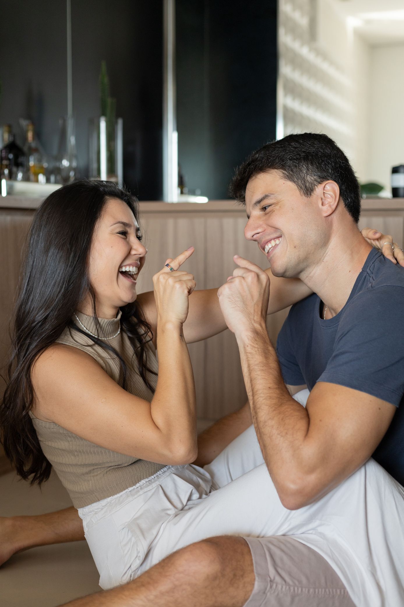 Happy couple laughing and pointing at each other playfully while sitting together in a modern kitchen.