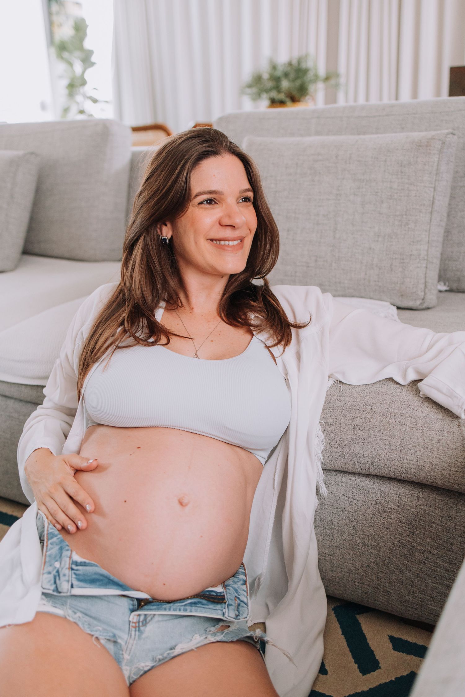 Expectant mother in casual white top and denim shorts relaxes on grey couch in bright living room.