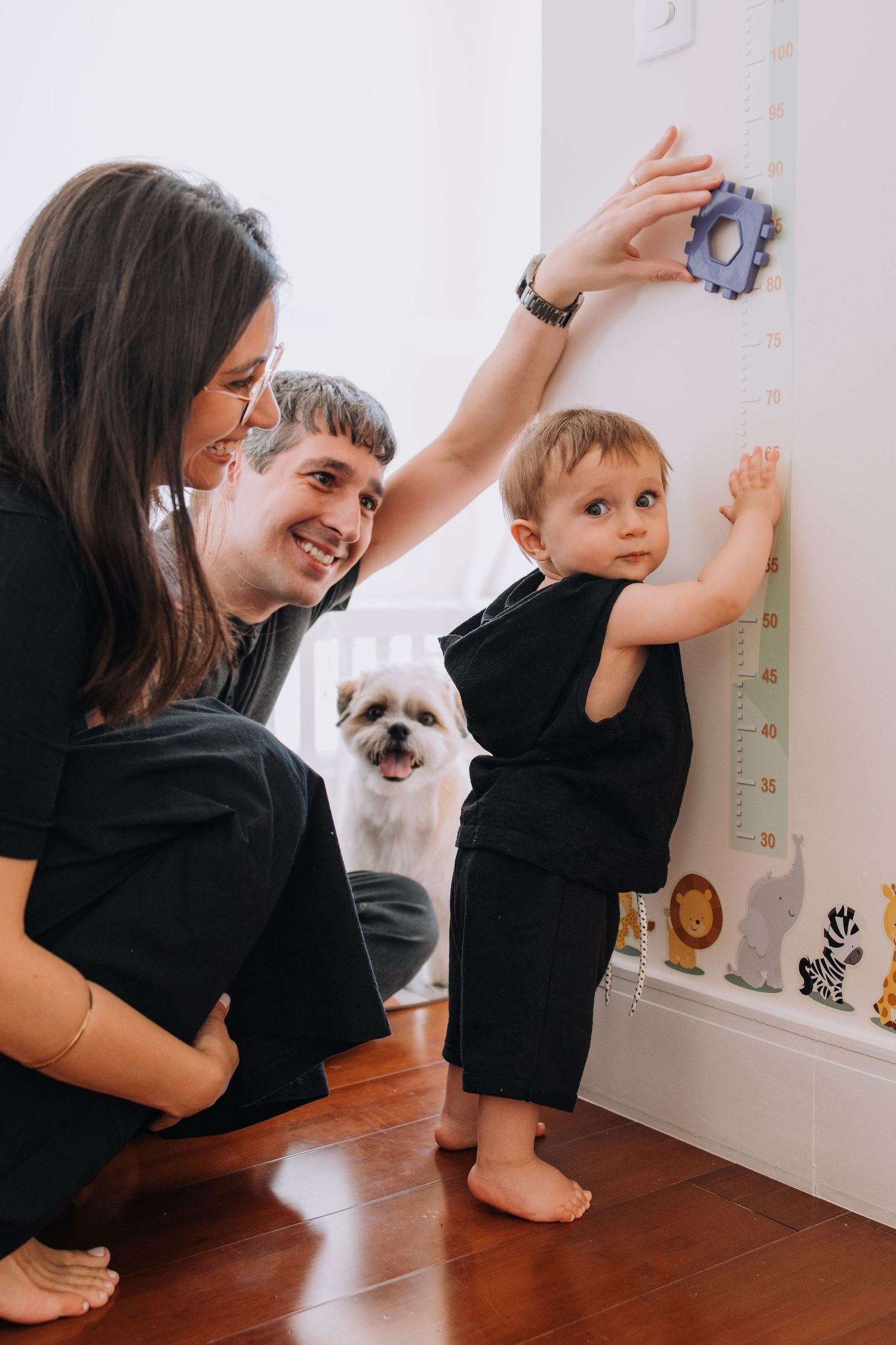 Family measuring child's height on wall growth chart with pet dog watching.