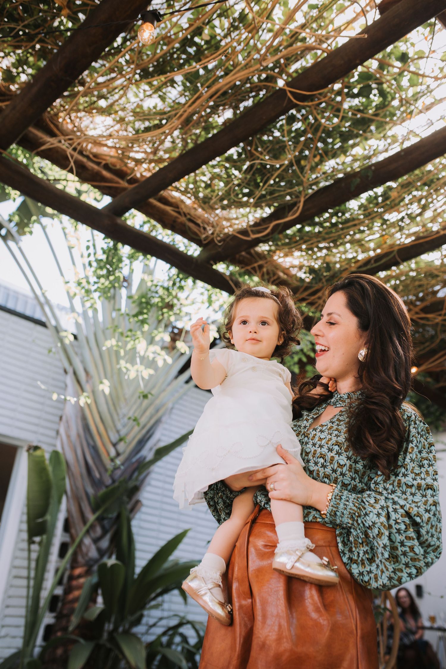 Mother and child sharing playful moment under vine-covered pergola.