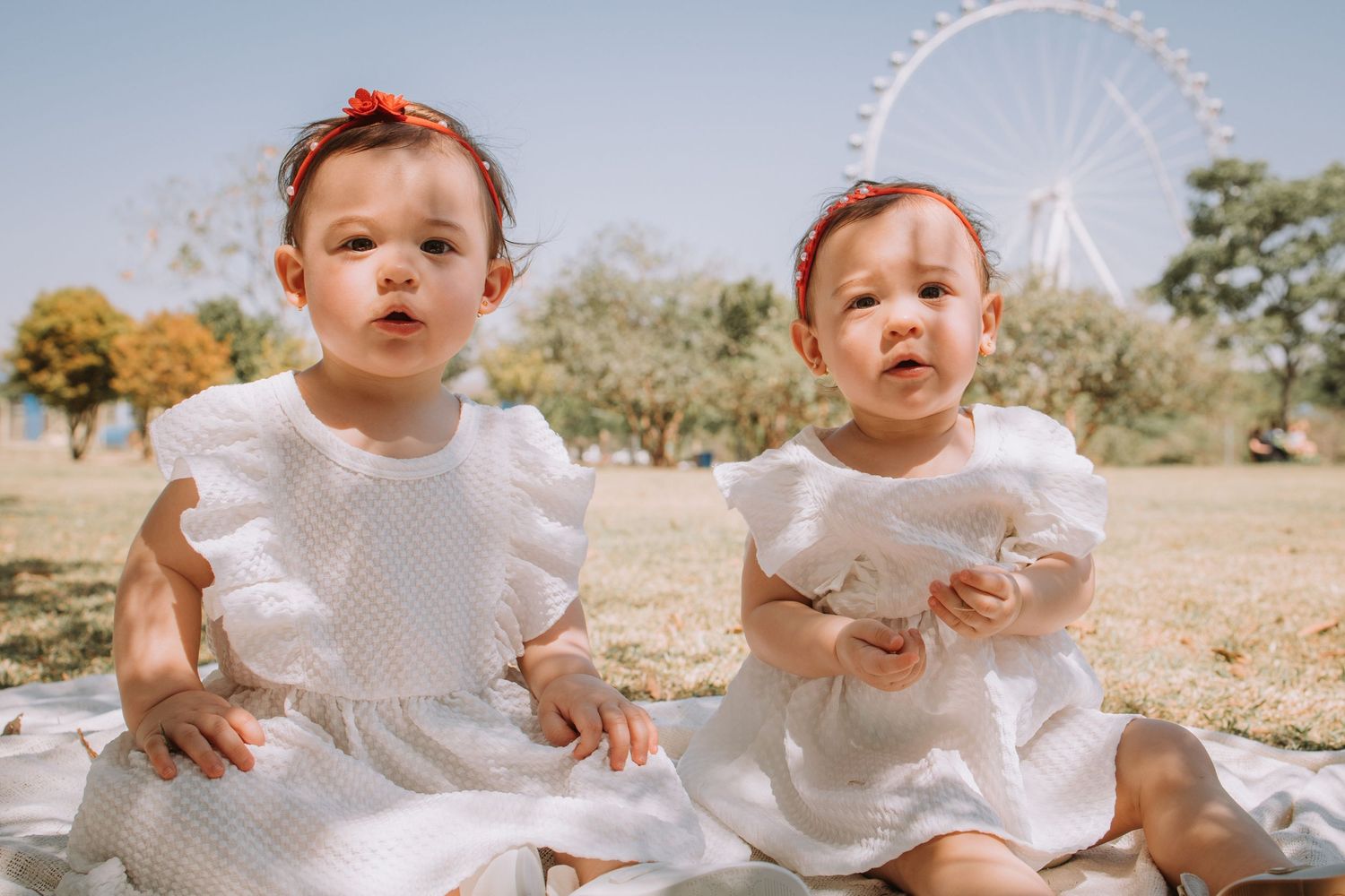 A baby in white dress sits on a blanket outdoors with trees and blue sky in the background.