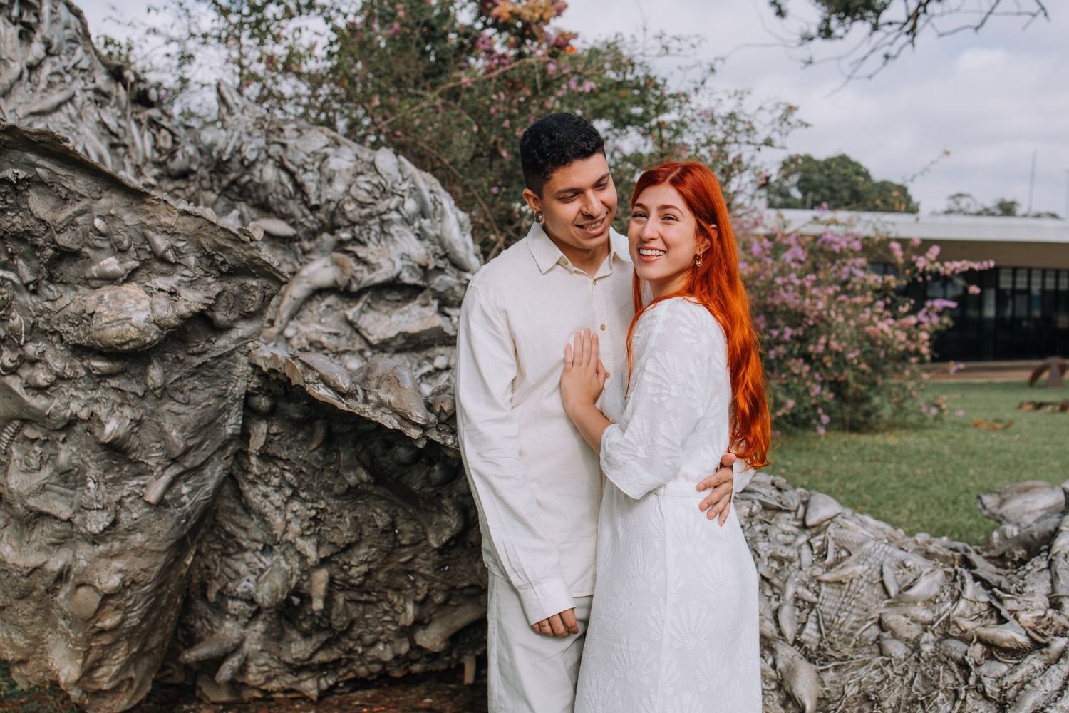 Couple in white outfits posing by a textured rock wall outdoors, woman with red hair smiling warmly.